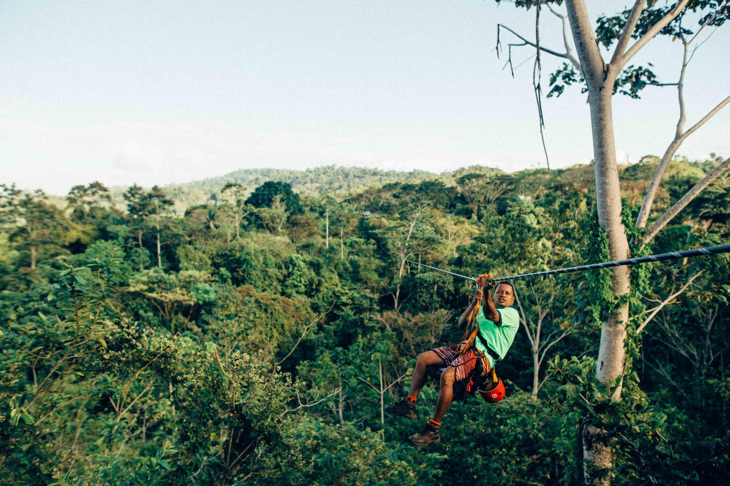 A person ziplines high above a dense, tropical jungle canopy with tree-covered mountains visible in the distance under a clear sky.