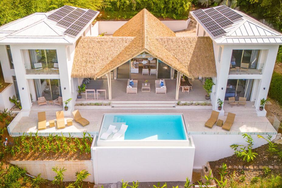 An aerial view showcases a modern villa with white two-story sections, traditional thatched roofing over an open central living area, and an infinity pool surrounded by a deck, all enveloped by dense tropical foliage.