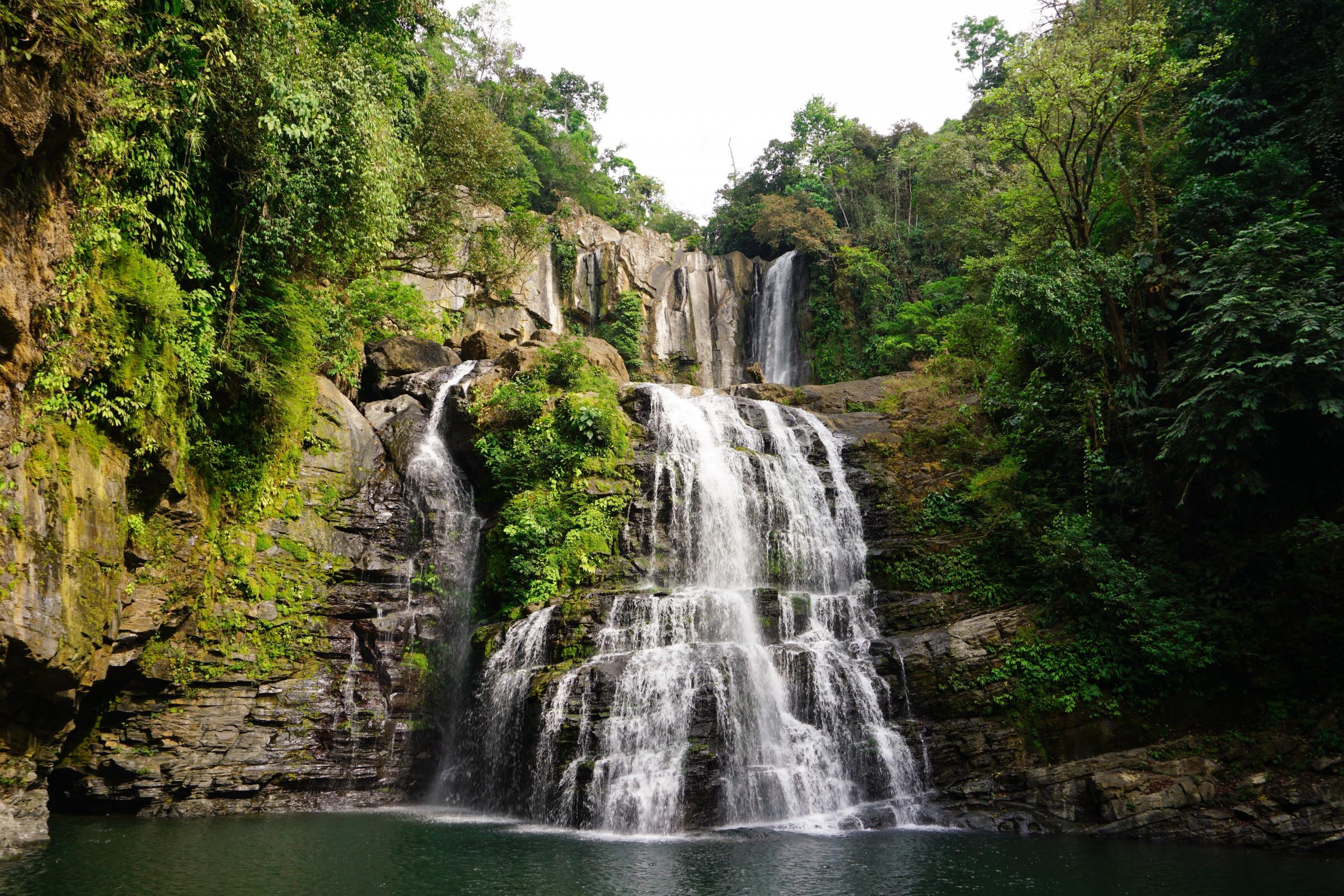 A multi-tiered waterfall cascades into a natural pool, surrounded by lush green jungle and rocky cliffs.