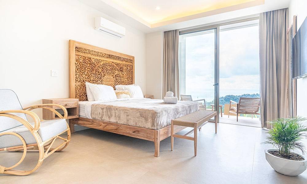 This bright bedroom features a bed with a carved wooden headboard, opening through glass doors to a balcony offering ocean and jungle views.