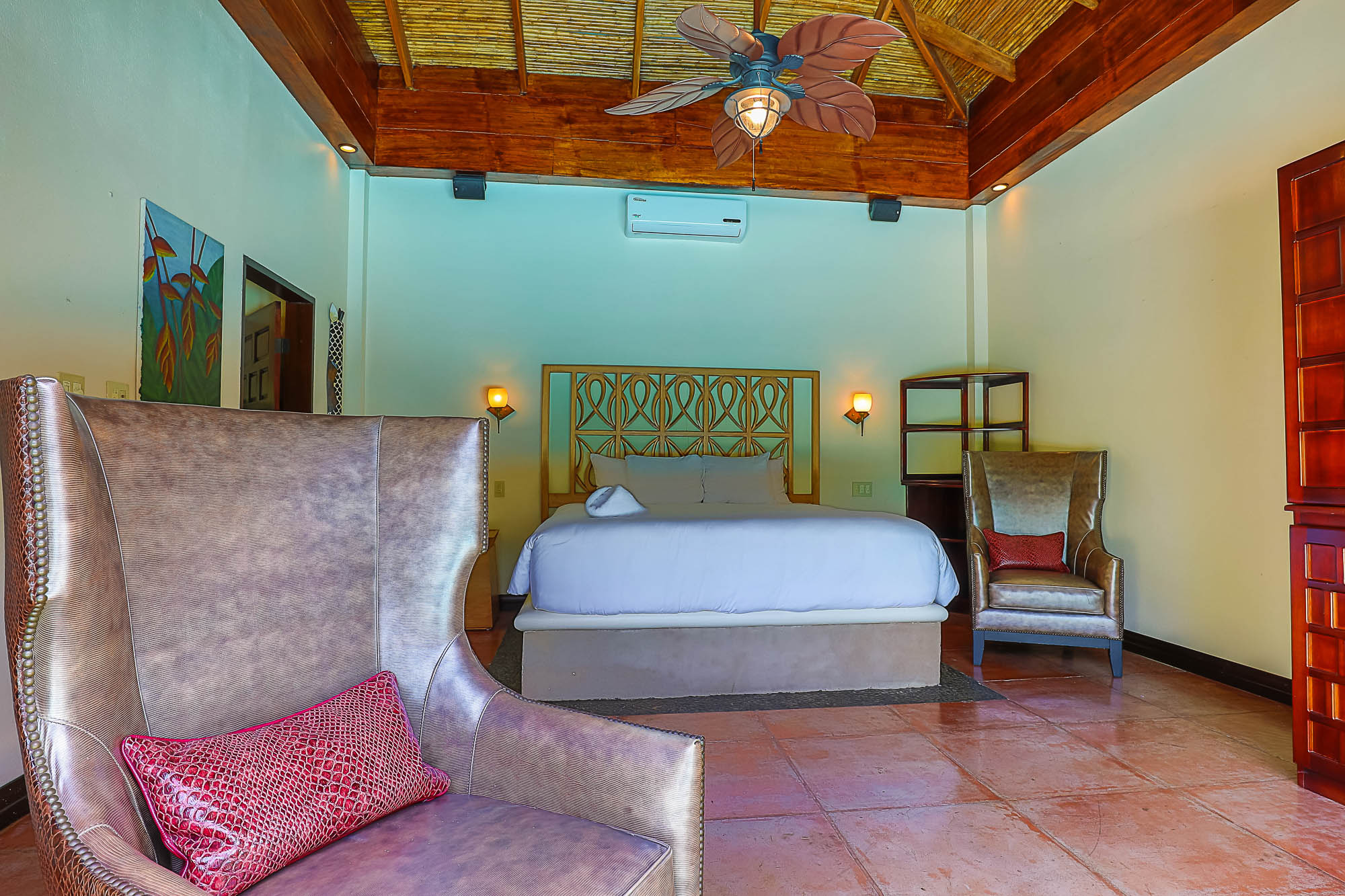 This inviting bedroom features a king bed with an ornate wooden headboard, a vaulted ceiling with exposed wooden beams and a tropical fan, and comfortable armchairs set on terracotta tile floors.