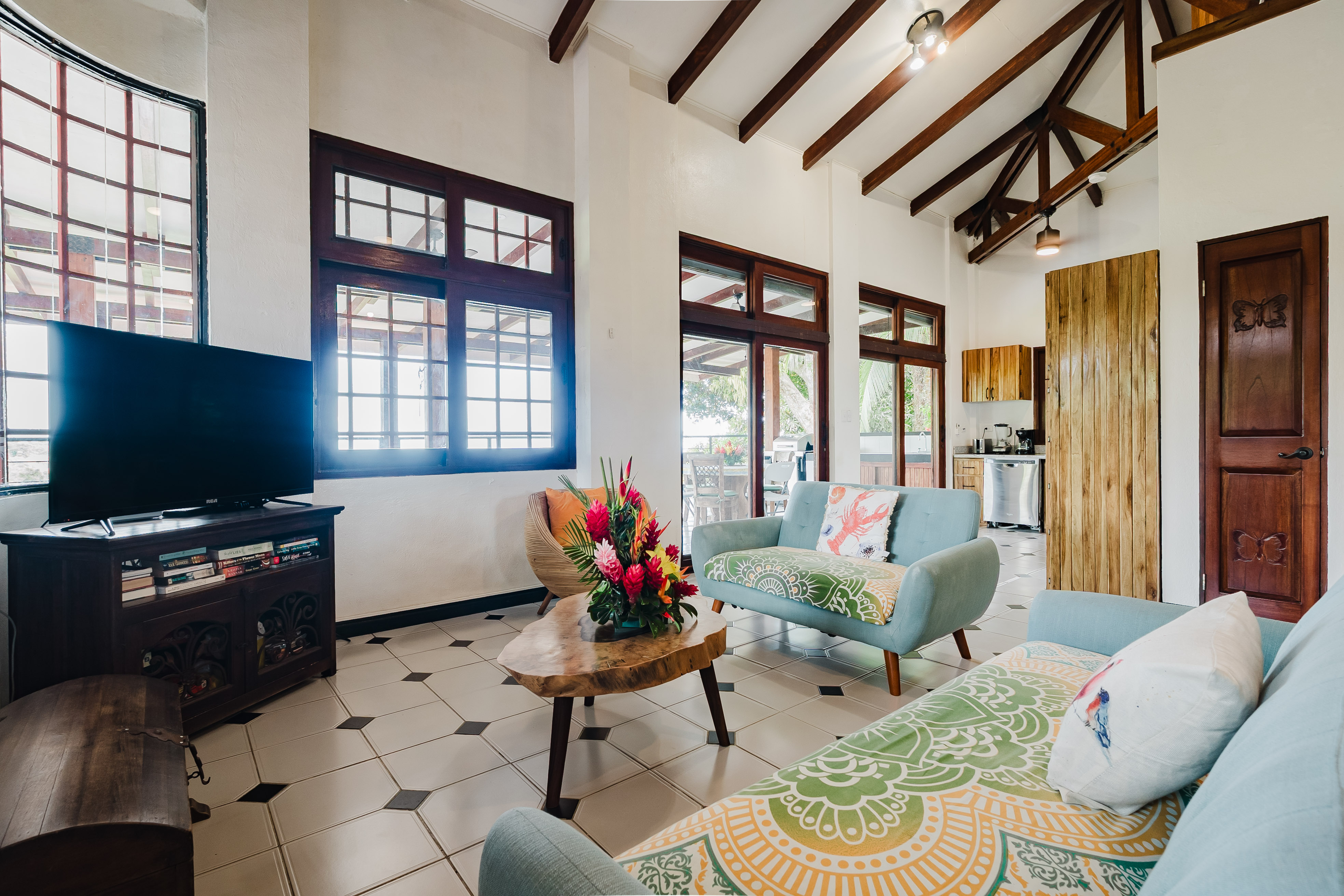 The bright living room features high ceilings with exposed wooden beams, large windows, and sliding glass doors that open to an outdoor terrace.
