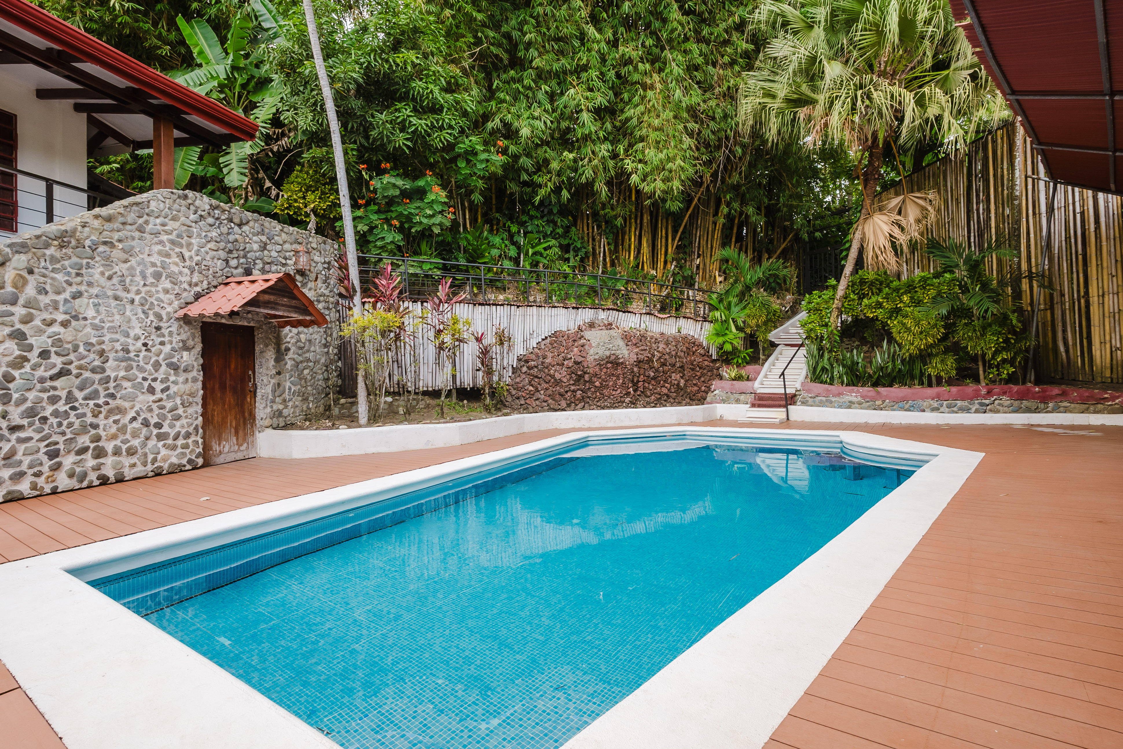 A blue-tiled swimming pool surrounded by a brown deck is set against a backdrop of lush tropical foliage and a stone-faced structure with a red-roofed balcony.