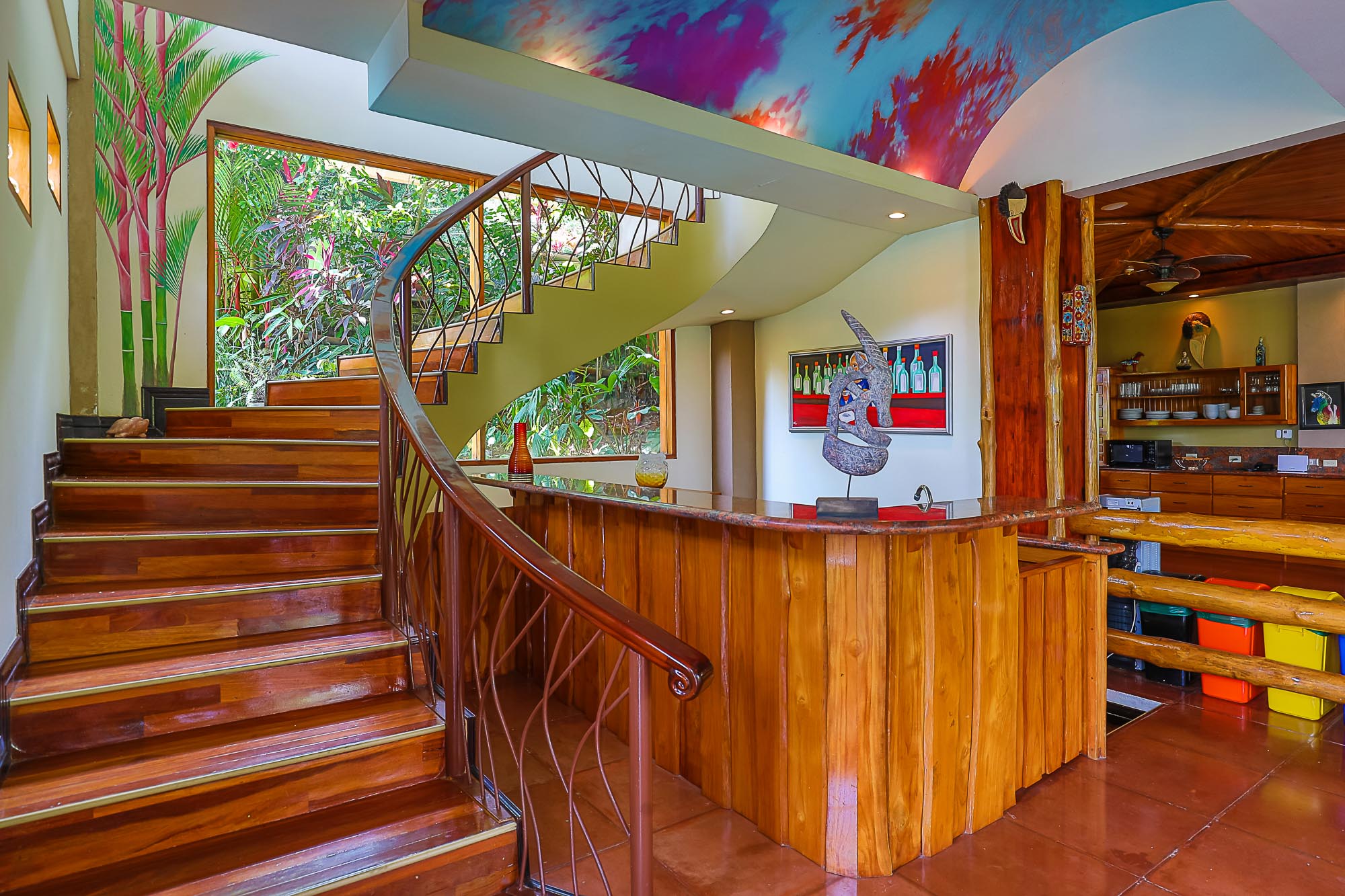 A grand wooden spiral staircase with an ornate metal railing ascends past large windows revealing lush tropical foliage, beneath a vibrant, mural-painted ceiling.