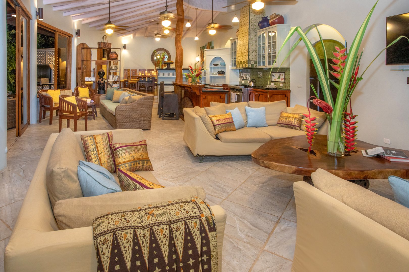 An expansive open-plan living space offers multiple comfortable seating areas, integrating a kitchen and a dining zone under high beamed ceilings, with large glass doors providing natural light and outdoor access.