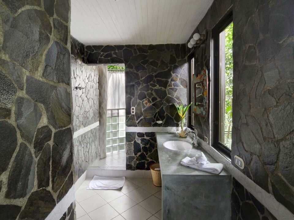 This spacious bathroom features dark stone walls, a double vanity with a concrete countertop, and large windows providing natural light and views of tropical foliage.
