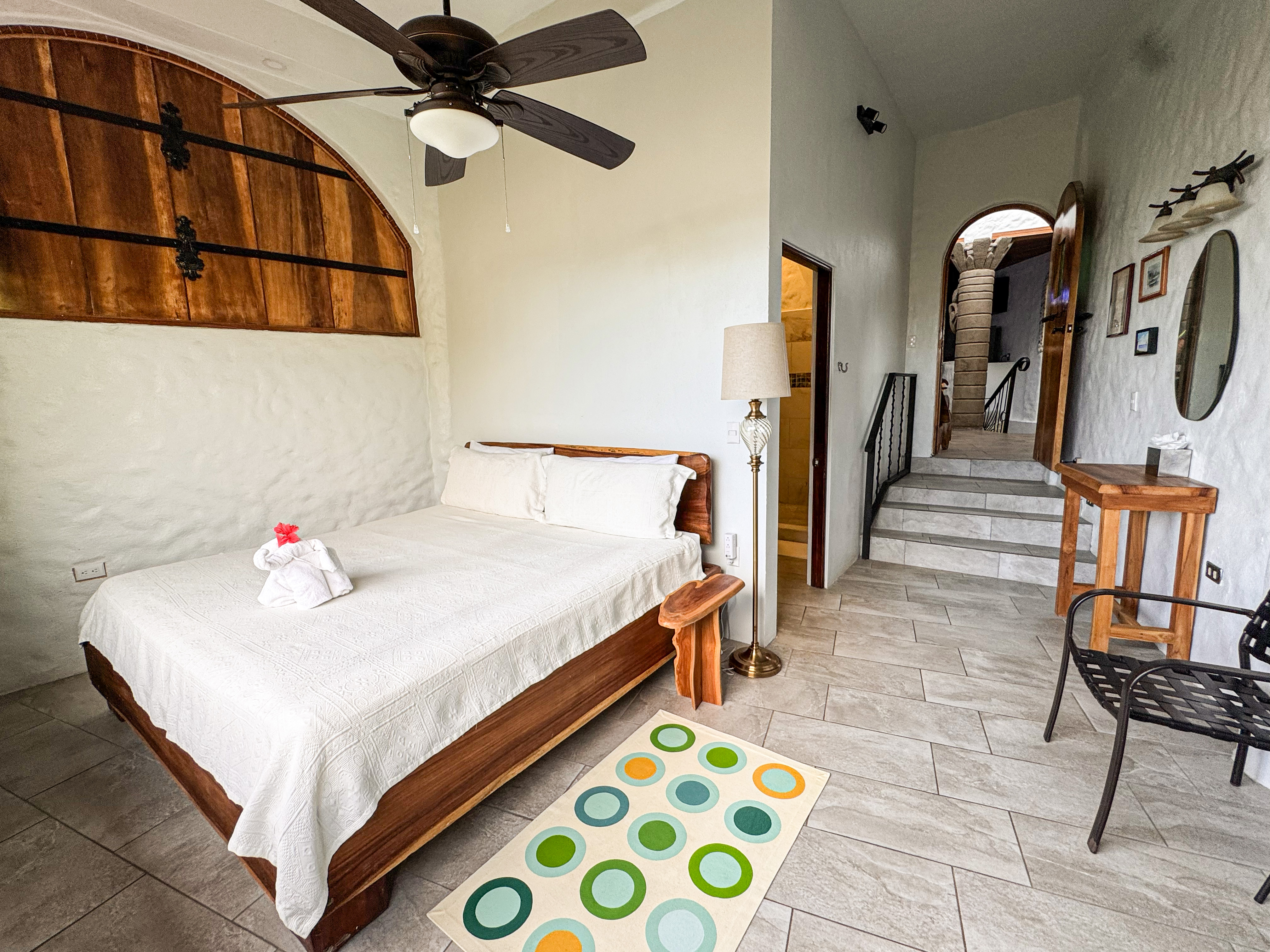 This bedroom features a wooden bed frame, textured white walls with an overhead arched timber panel, and bright illumination, connecting to a private bathroom and upper level via stairs.