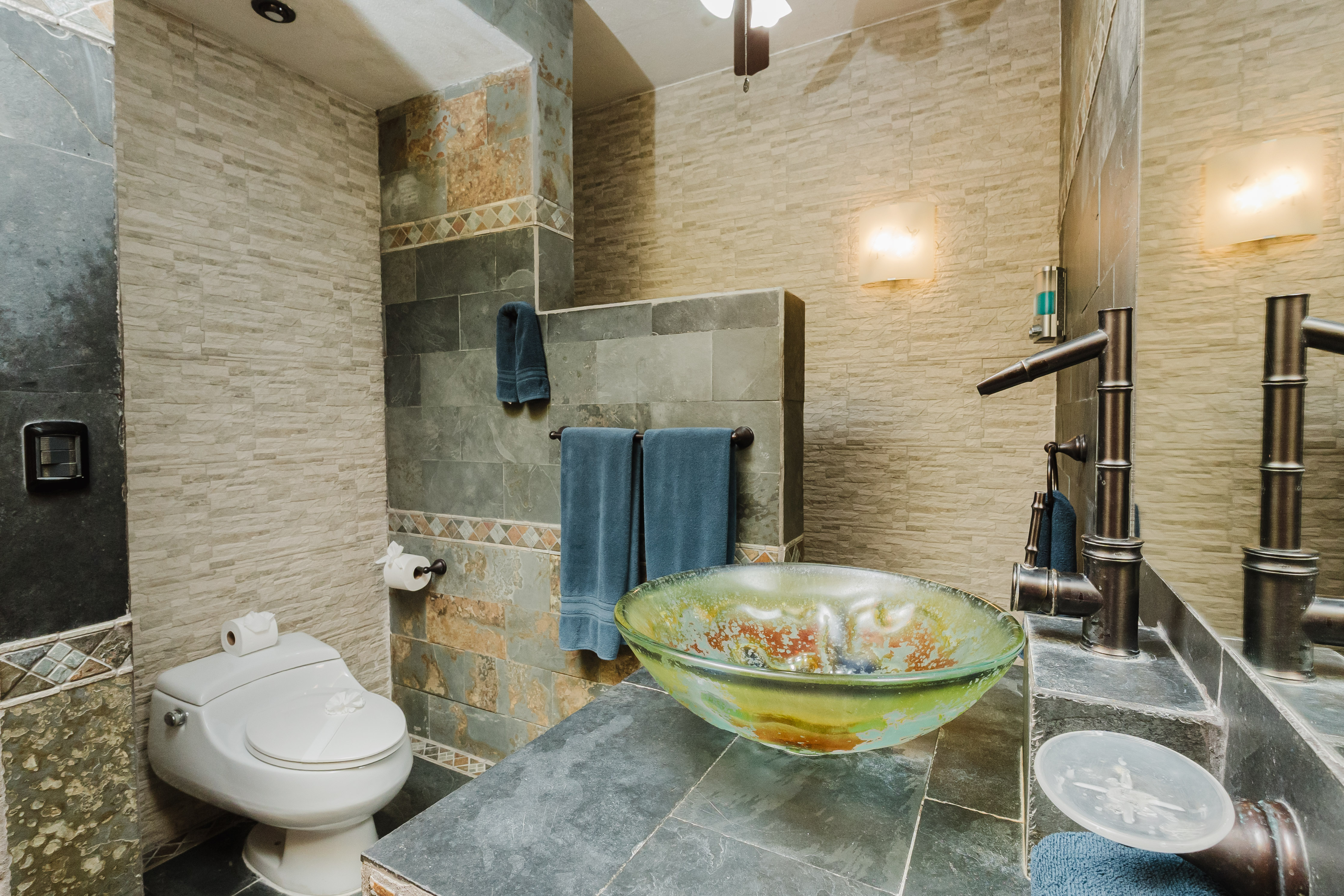 A bathroom features textured stone and tile walls, a dark slate vanity with a distinctive glass vessel sink, a bamboo-style bronze faucet, and a toilet.