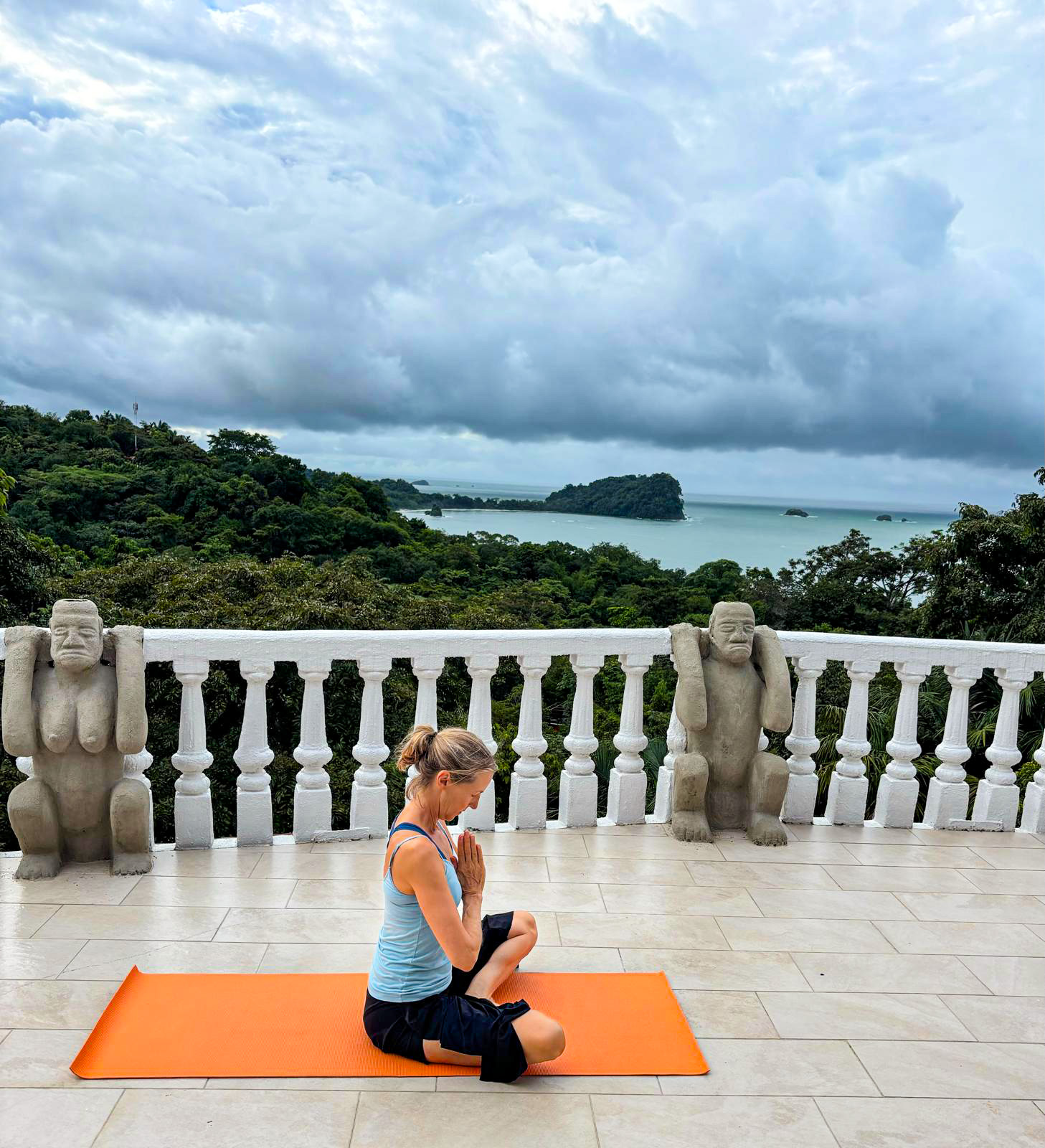 An outdoor tiled terrace features a white balustrade and two carved stone figures, overlooking a tropical jungle canopy and the Pacific Ocean.