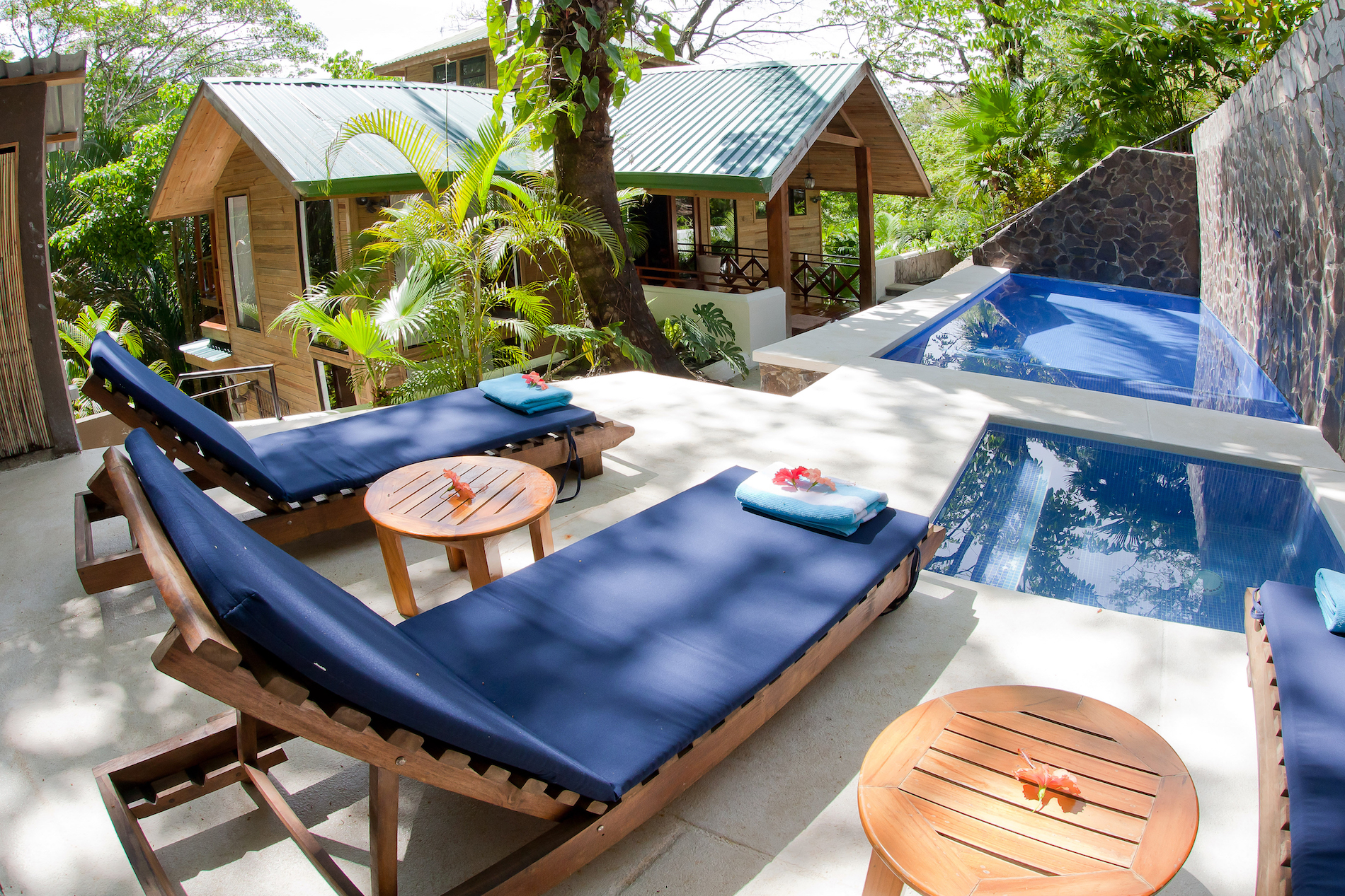 A sunlit terrace with lounge chairs and a private pool and jacuzzi provides views of the wooden villa and lush tropical jungle.
