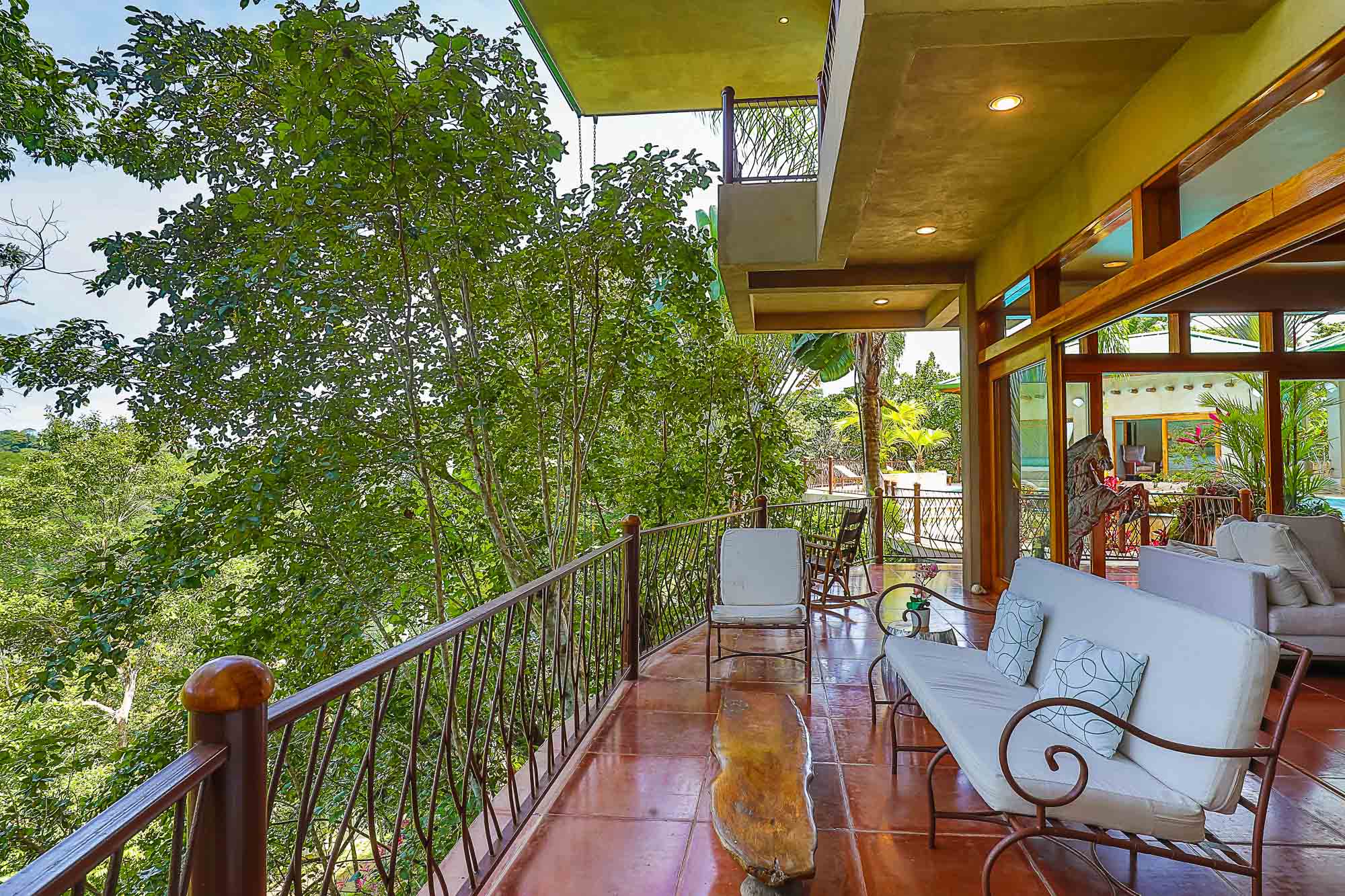 This outdoor terrace, featuring varied seating and natural wood elements, overlooks a vibrant rainforest canopy and opens into the villa's interior through wide glass doors.