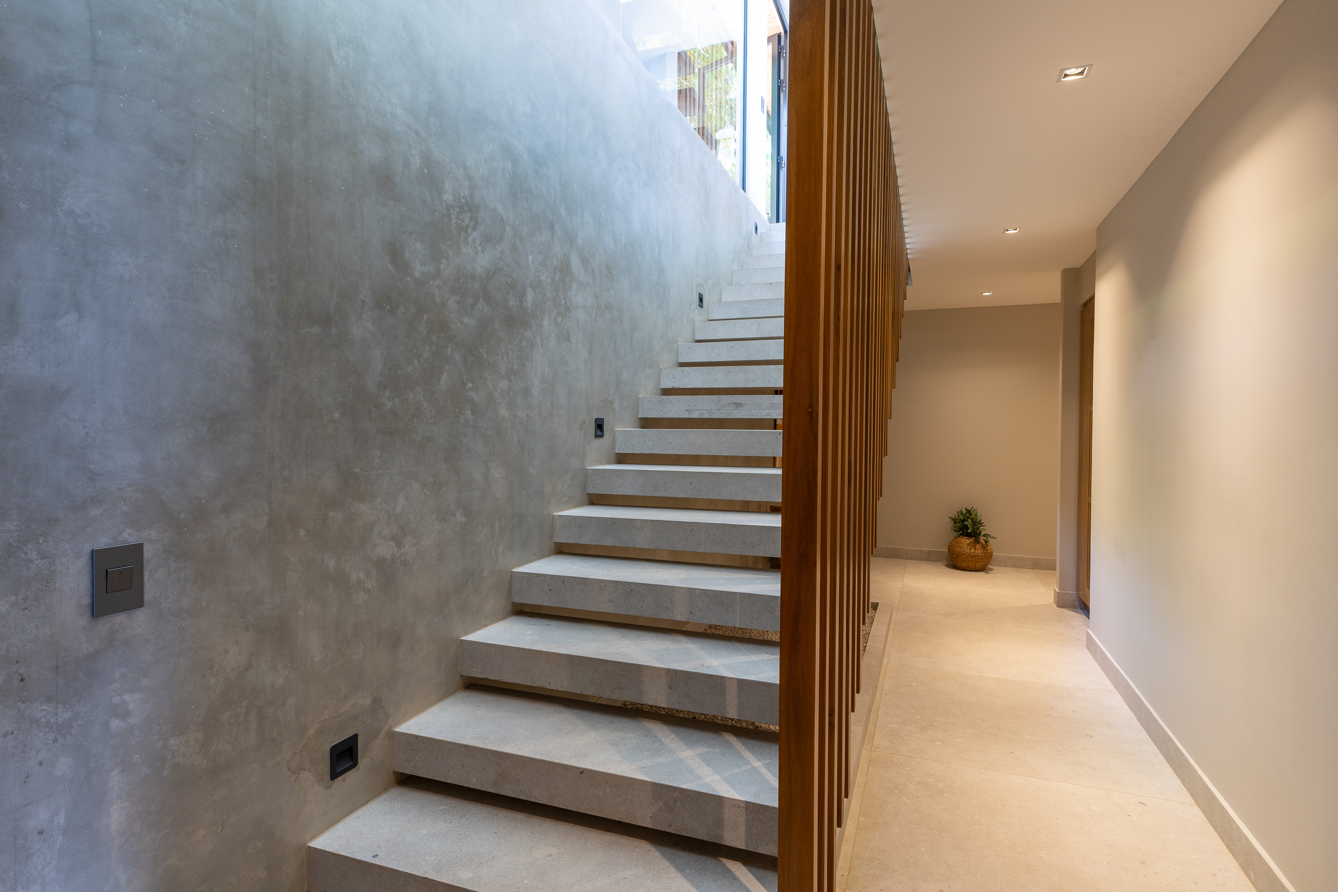 This contemporary interior staircase, featuring light concrete steps and a wooden slatted railing, is bathed in natural light from a large window overlooking the surrounding jungle.