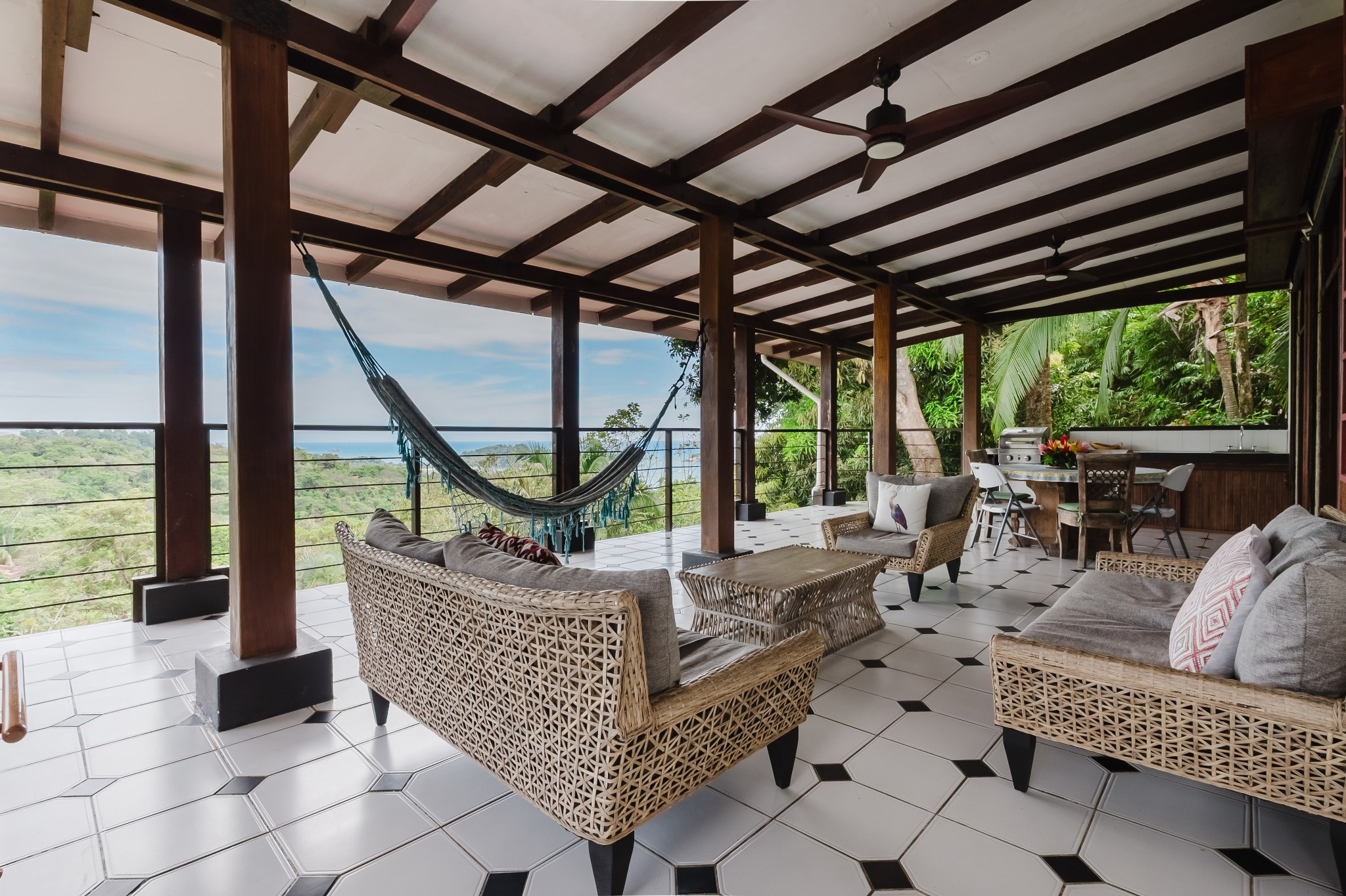 This open-air terrace features a beamed ceiling, tiled floor, various seating arrangements, a hammock, and an outdoor kitchen, all overlooking panoramic jungle and ocean views.