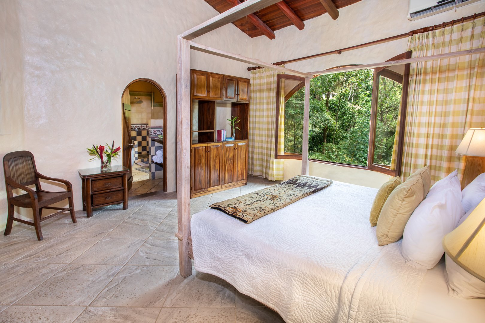 This bedroom offers a four-poster bed, a wooden beamed ceiling, and a large window providing a direct view into the tropical jungle, with an arched entry to a private bathroom.
