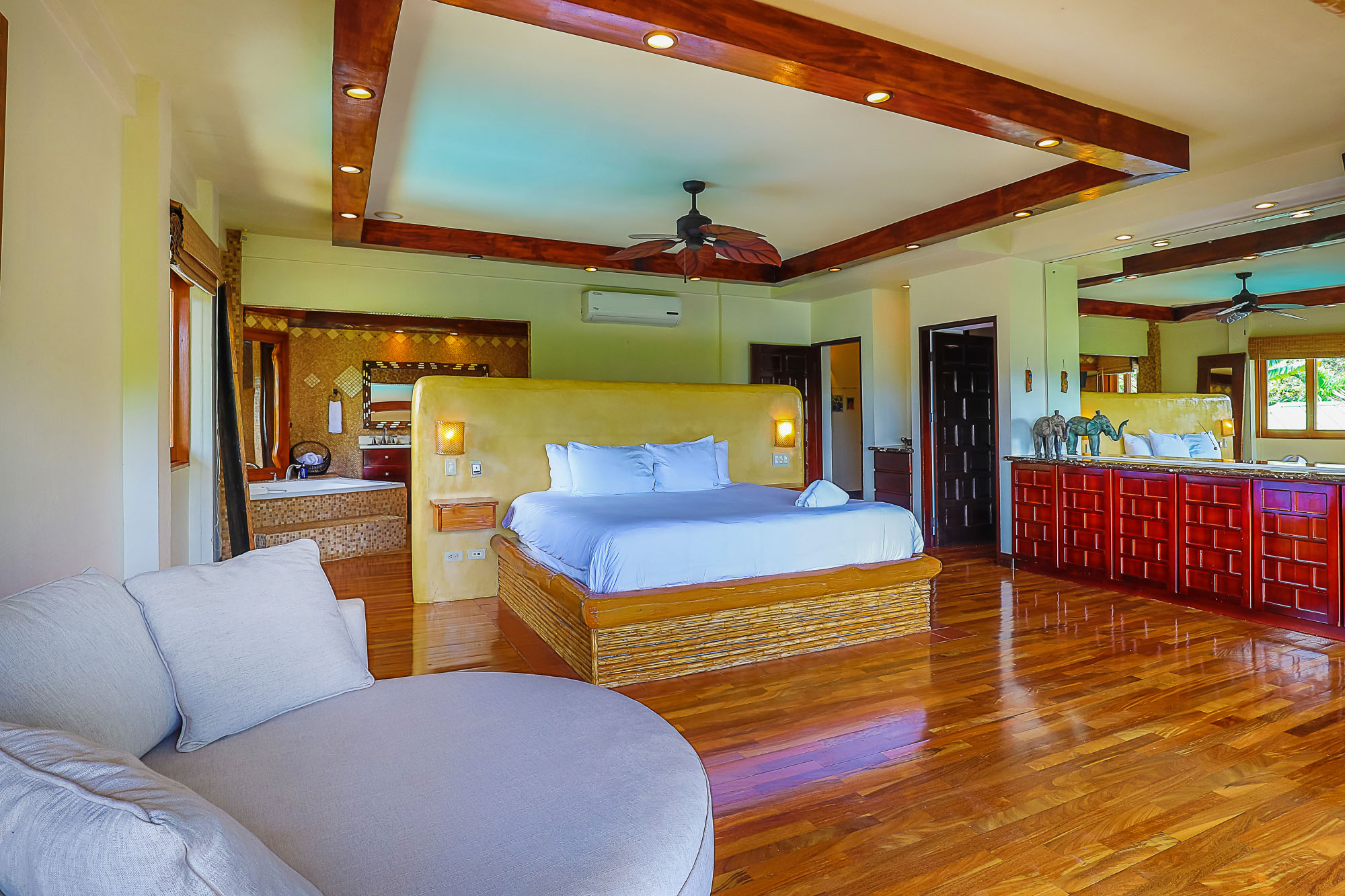 This spacious bedroom suite features a king bed with a bamboo-style base, polished wood floors, and a decorative tray ceiling with dark wood accents, offering natural light, tropical views, and an open-concept bathroom area with a soaking tub.