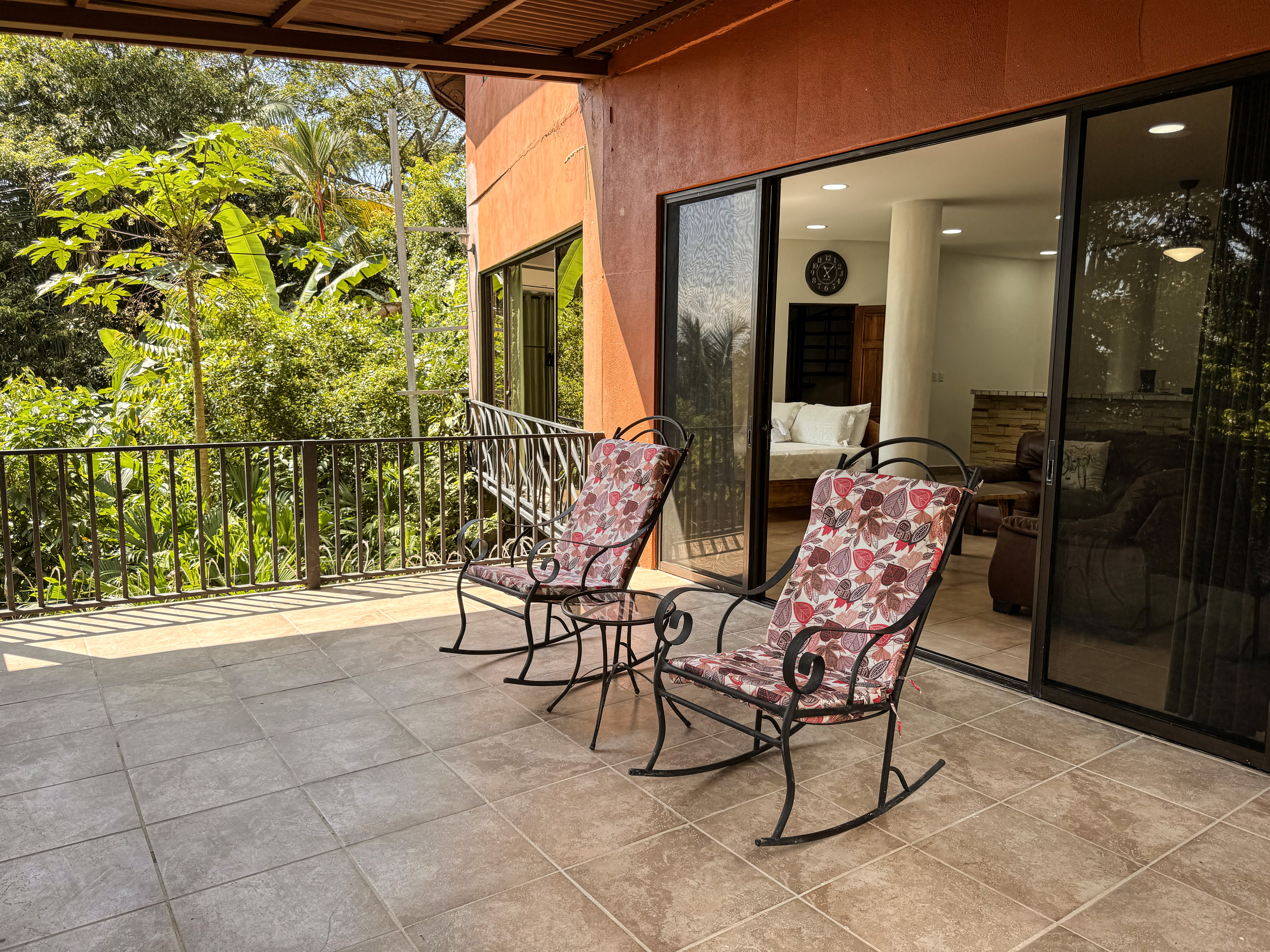 This sunlit tiled terrace, featuring a dark metal railing and partial roof, offers jungle views and connects directly to a bright bedroom through sliding glass doors.