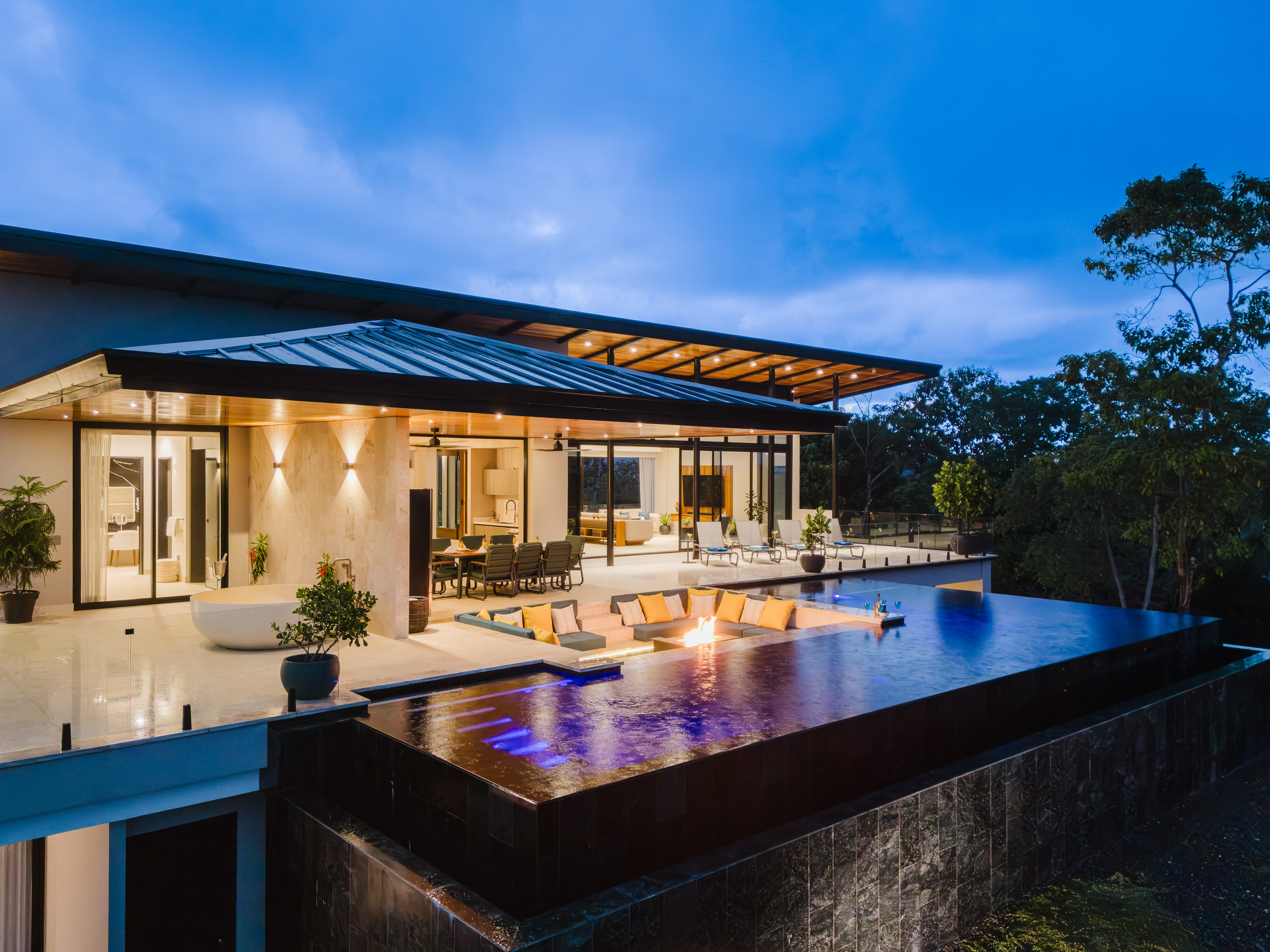 This modern villa at dusk features illuminated indoor living spaces opening onto a multi-level infinity pool and terrace, set against lush tropical vegetation.
