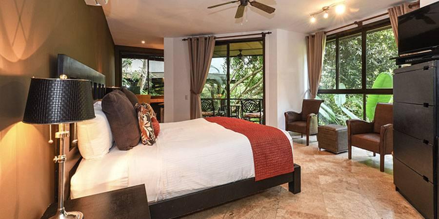 A bright bedroom features a large bed, a seating area, and a private balcony offering views of lush tropical foliage through sliding glass doors.