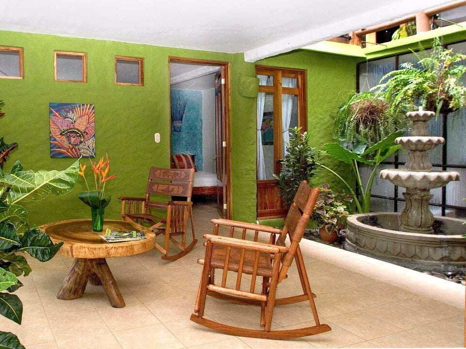 A private terrace, featuring vibrant green walls and natural wood rocking chairs, connects to a bedroom suite and overlooks a garden with a multi-tiered stone fountain.