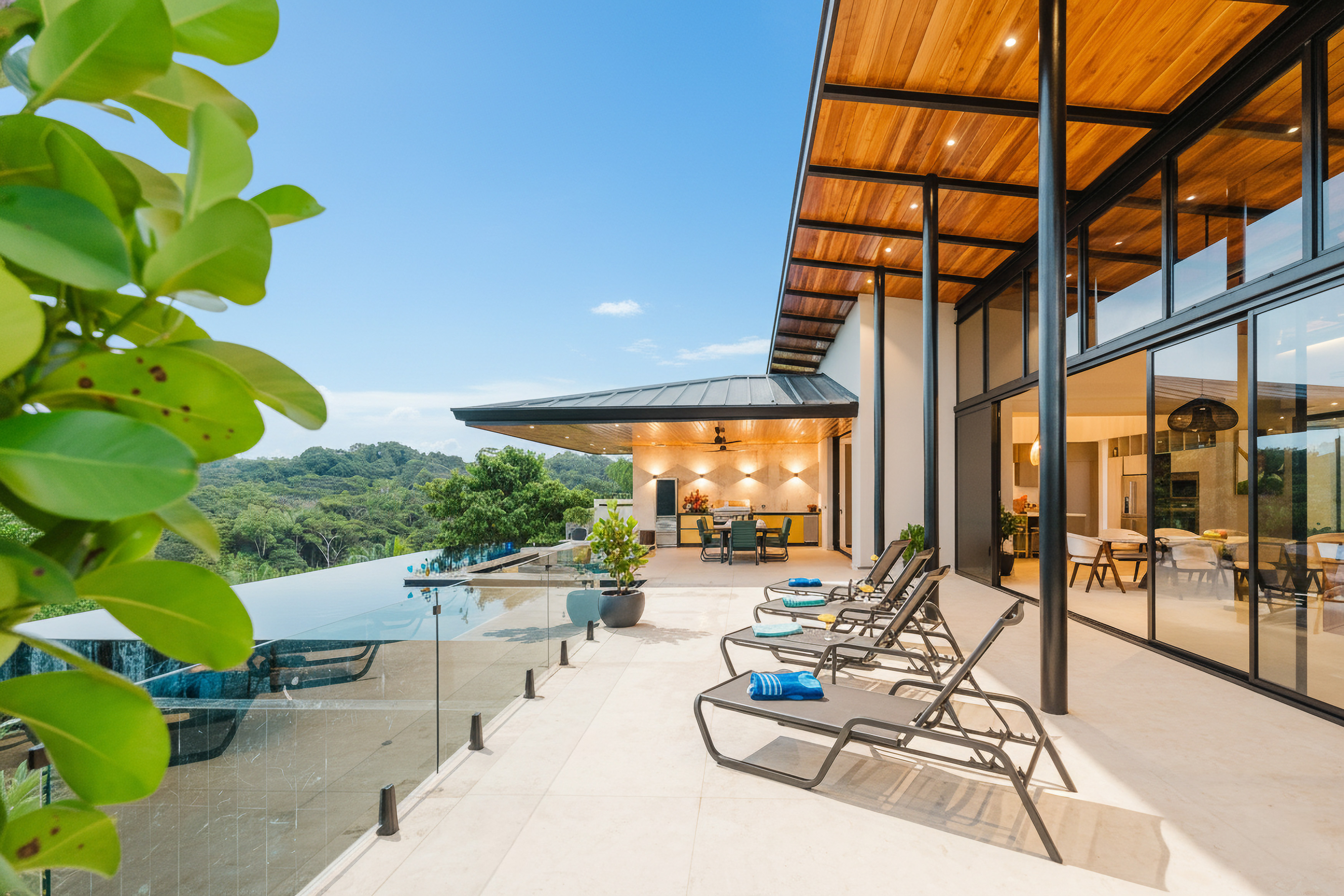 An open, sunlit terrace features an infinity pool with glass railings, an outdoor dining area, and unobstructed views of the jungle, connecting to the villa's modern interior through tall glass doors.