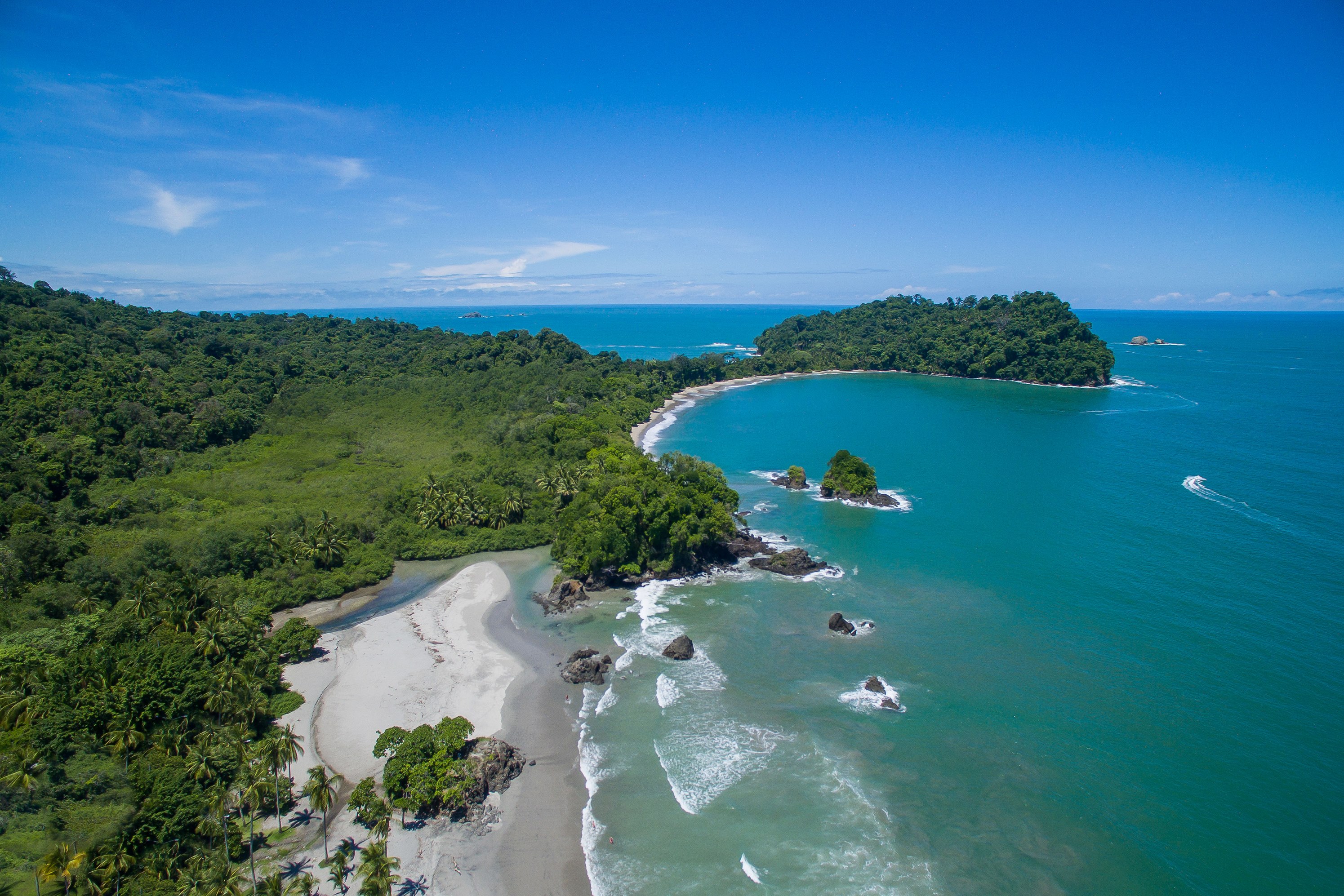 An aerial view showcases the vast Pacific Ocean meeting a curving sandy beach and dense tropical jungle, dotted with rocky islets under a clear, bright sky.