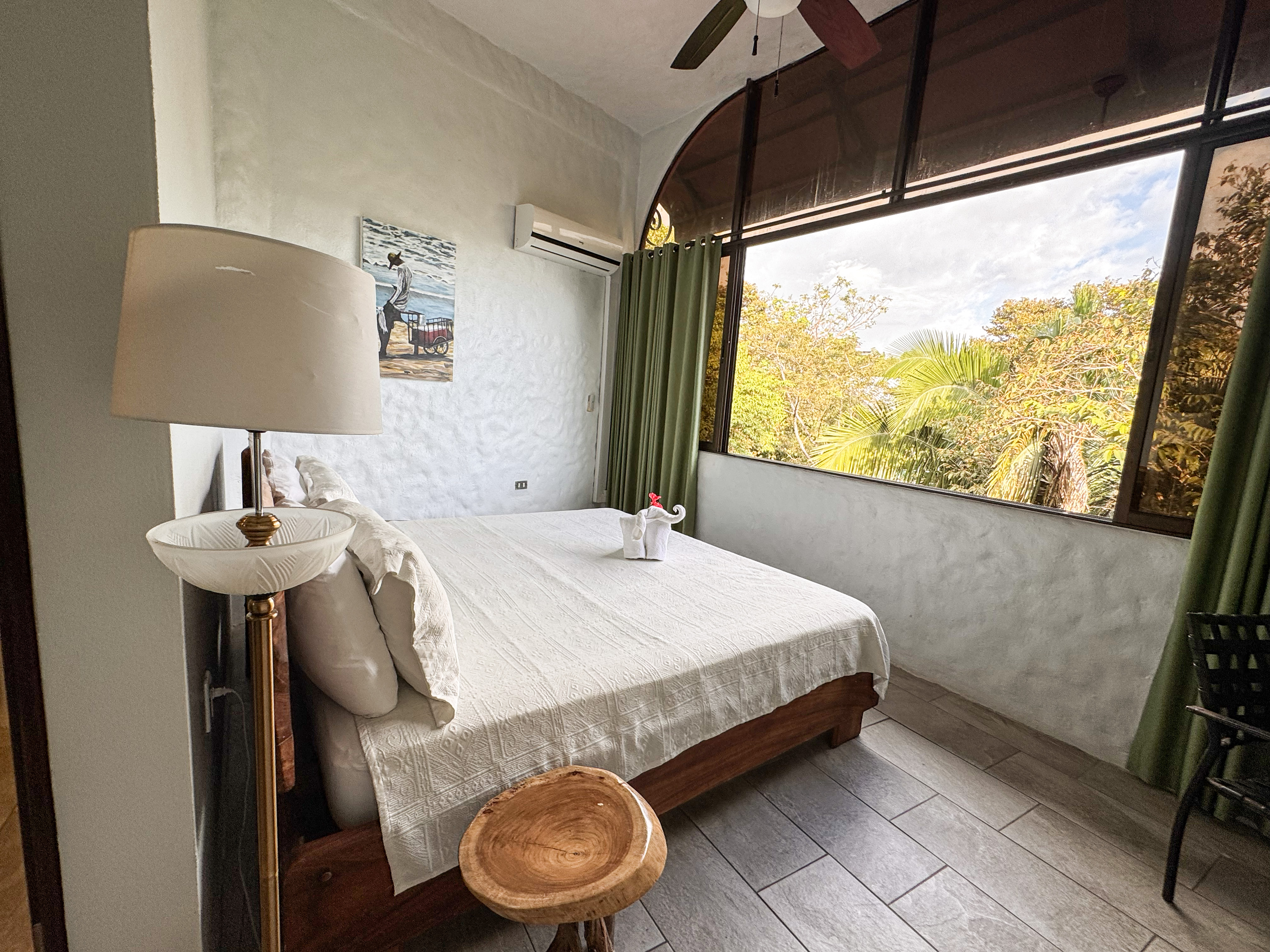 This bedroom features a comfortable bed, textured walls, and a large arched window framing tropical jungle views, allowing abundant natural light.
