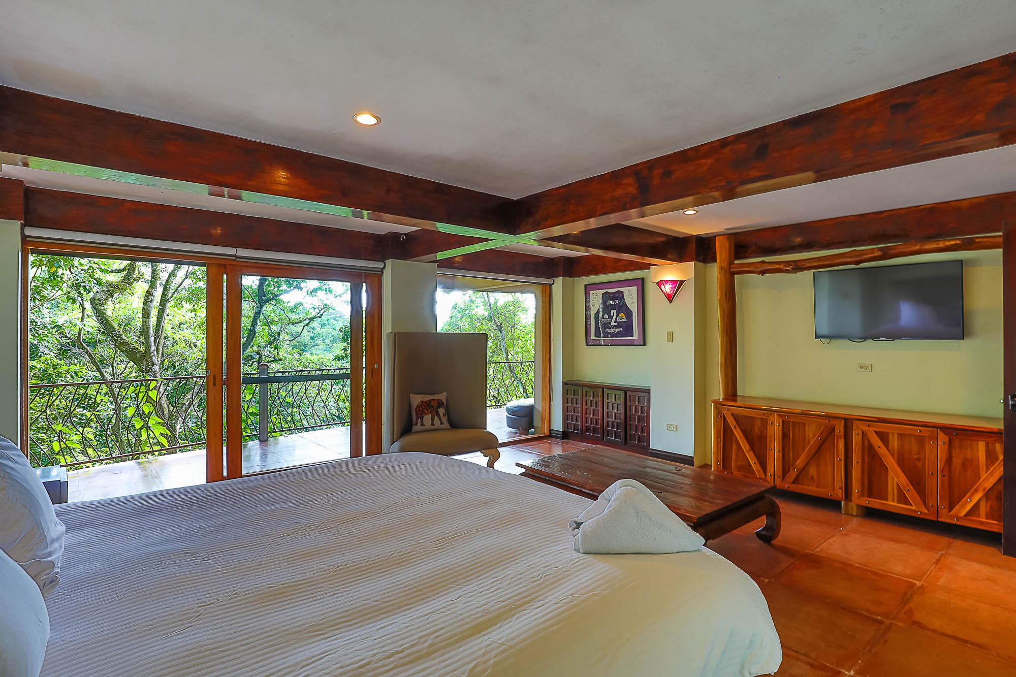 This bedroom features exposed wooden beams, a spacious bed, and expansive glass doors opening to a private terrace with direct views of the surrounding rainforest, bathing the room in natural light.