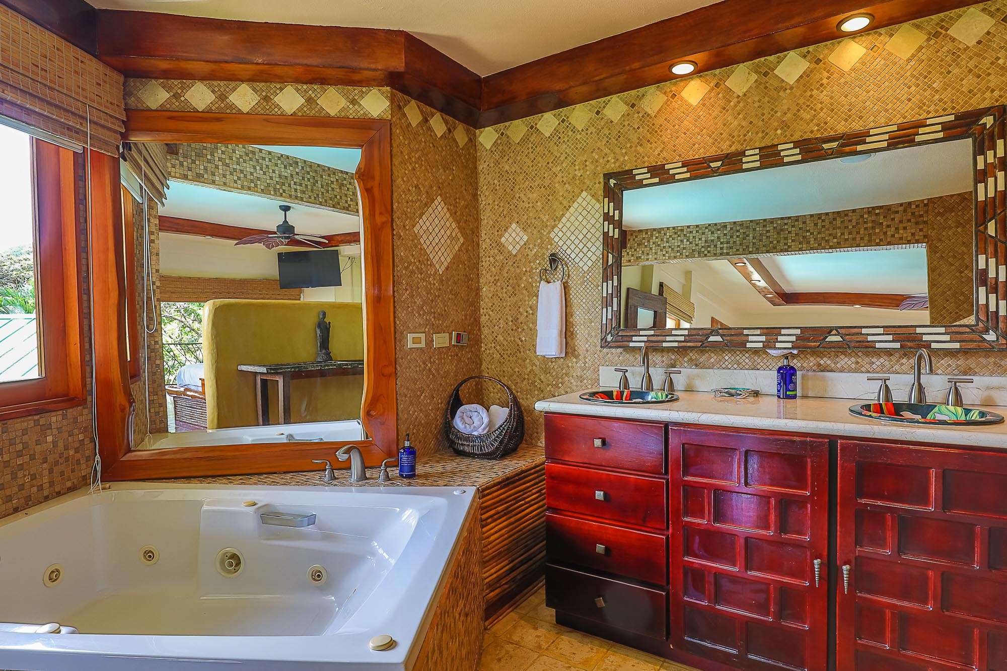 A spacious bathroom features a jetted tub, a double vanity with dark wood cabinetry, and mosaic-tiled walls, with a large window providing natural light and views of tropical foliage.