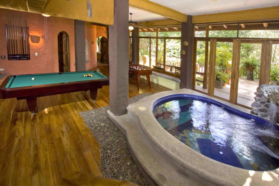 This game lounge features a full-size pool table, foosball, and an indoor splash pool with a waterfall, surrounded by large glass doors that provide natural light and views of the tropical greenery.