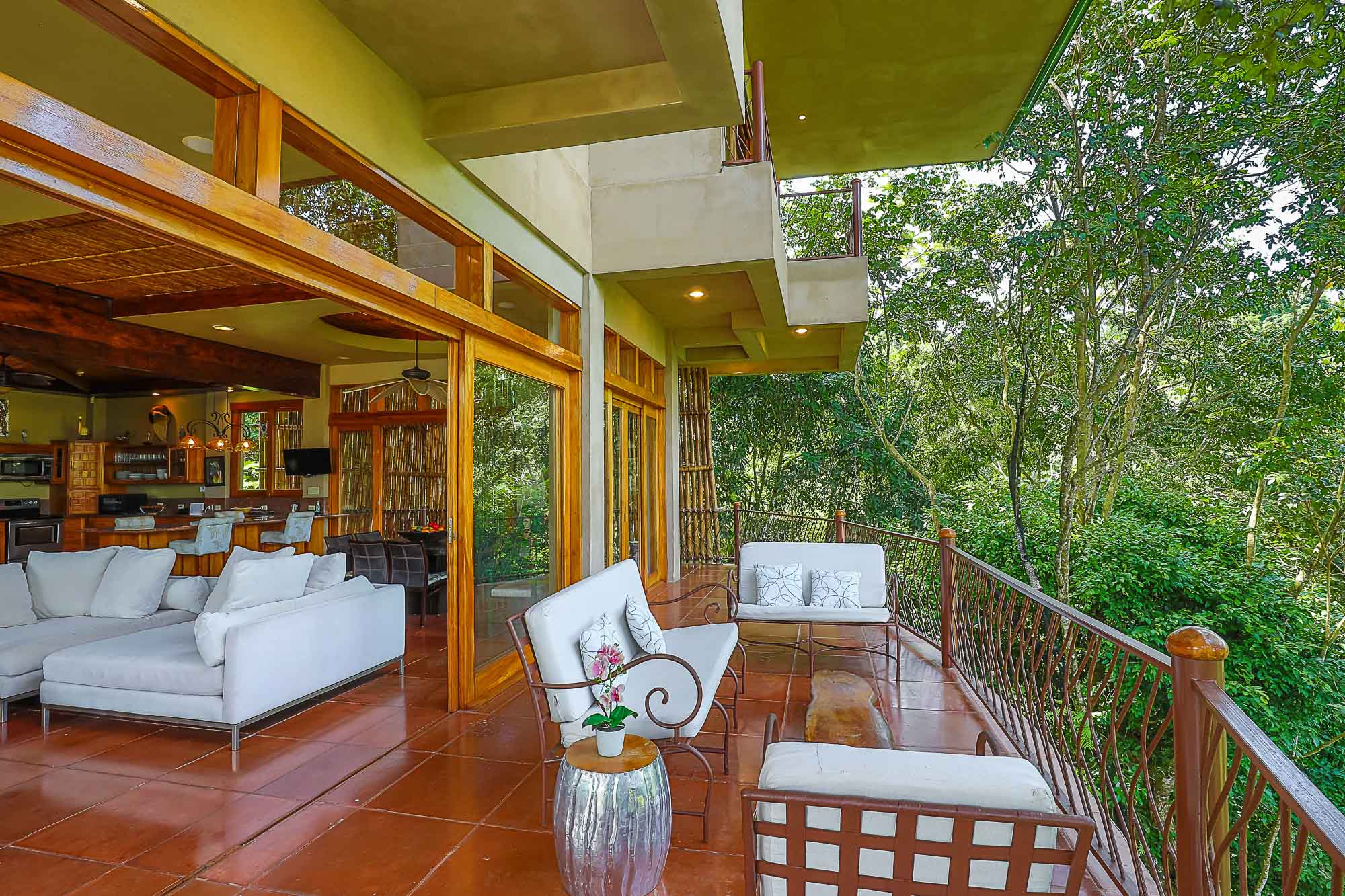 The villa's main level terrace features an open-air seating area with direct views into the tropical rainforest, connected to the interior living room and kitchen by wide wooden-framed glass doors.