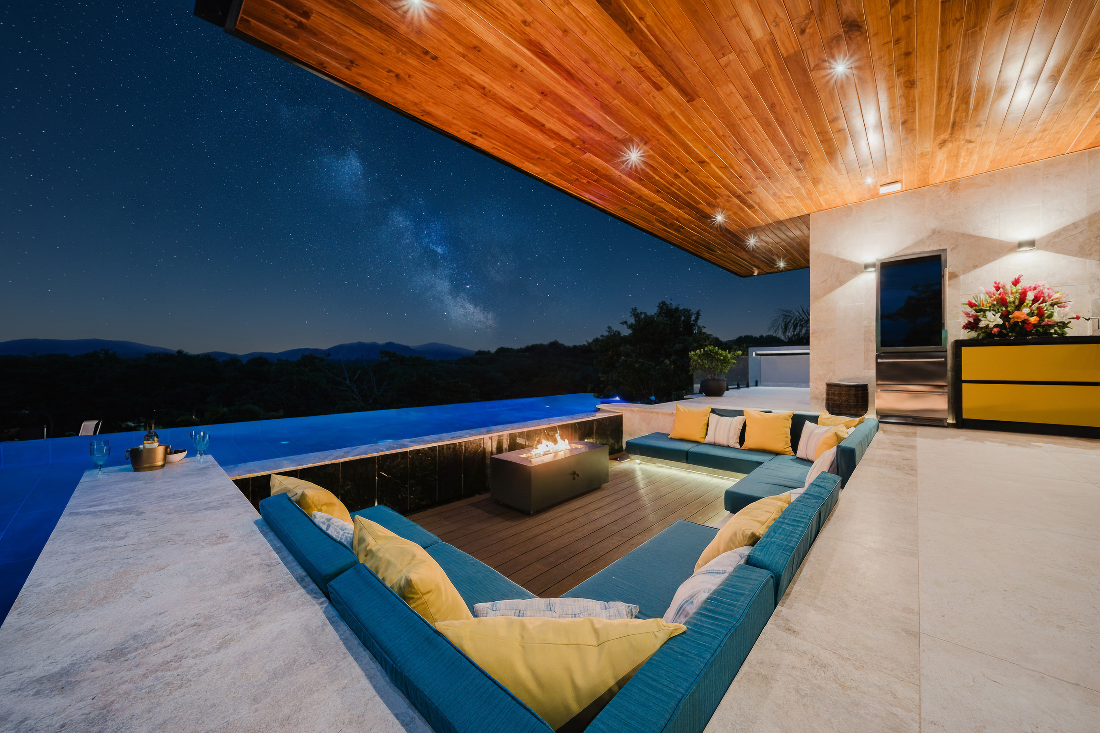 A covered outdoor terrace with an L-shaped lounge and fire pit overlooks an infinity pool and a night sky filled with stars above distant mountains.