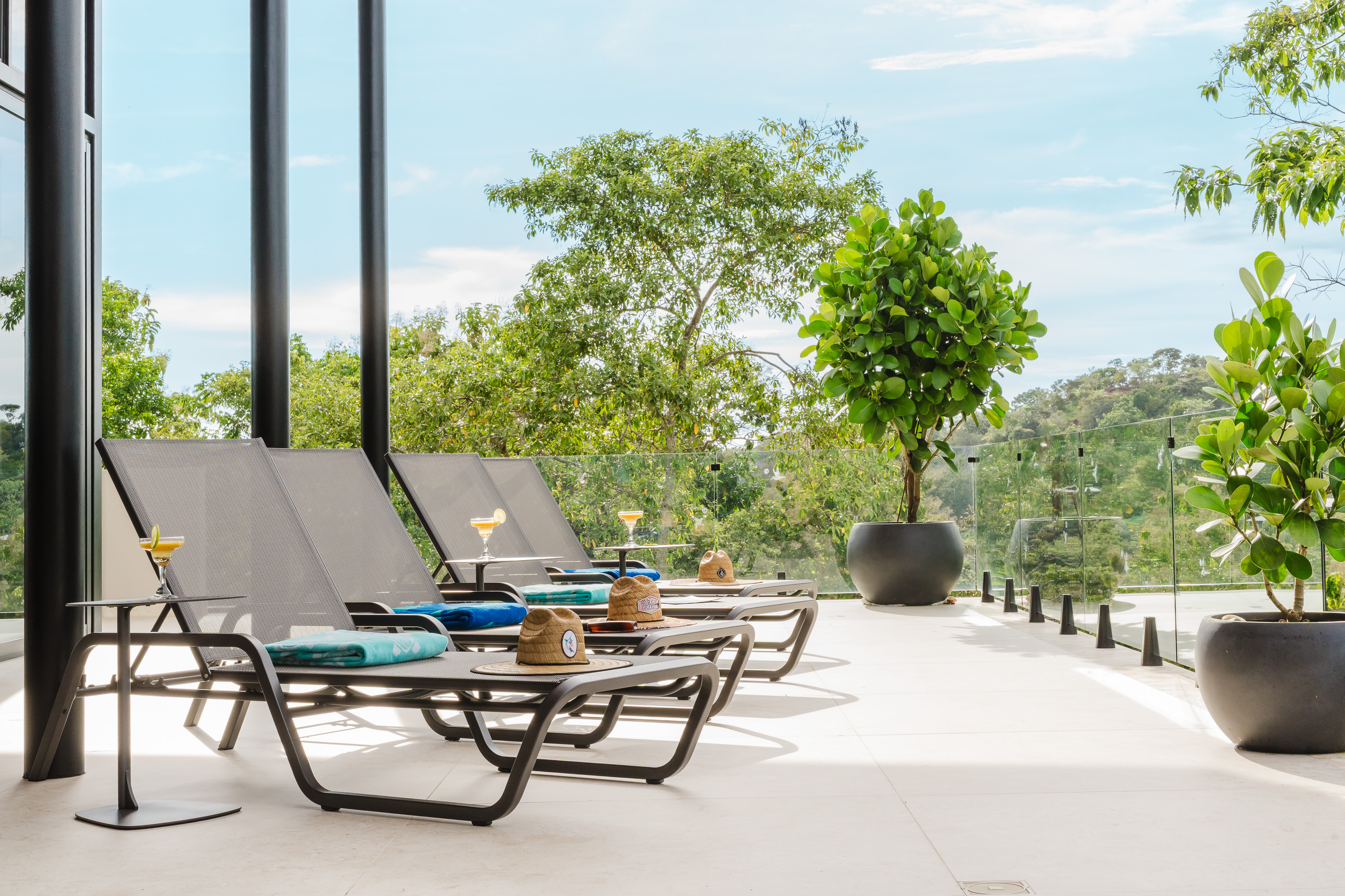 A bright outdoor terrace features lounge chairs on light-colored tile, opening to expansive tropical tree views and the sky through a glass railing.