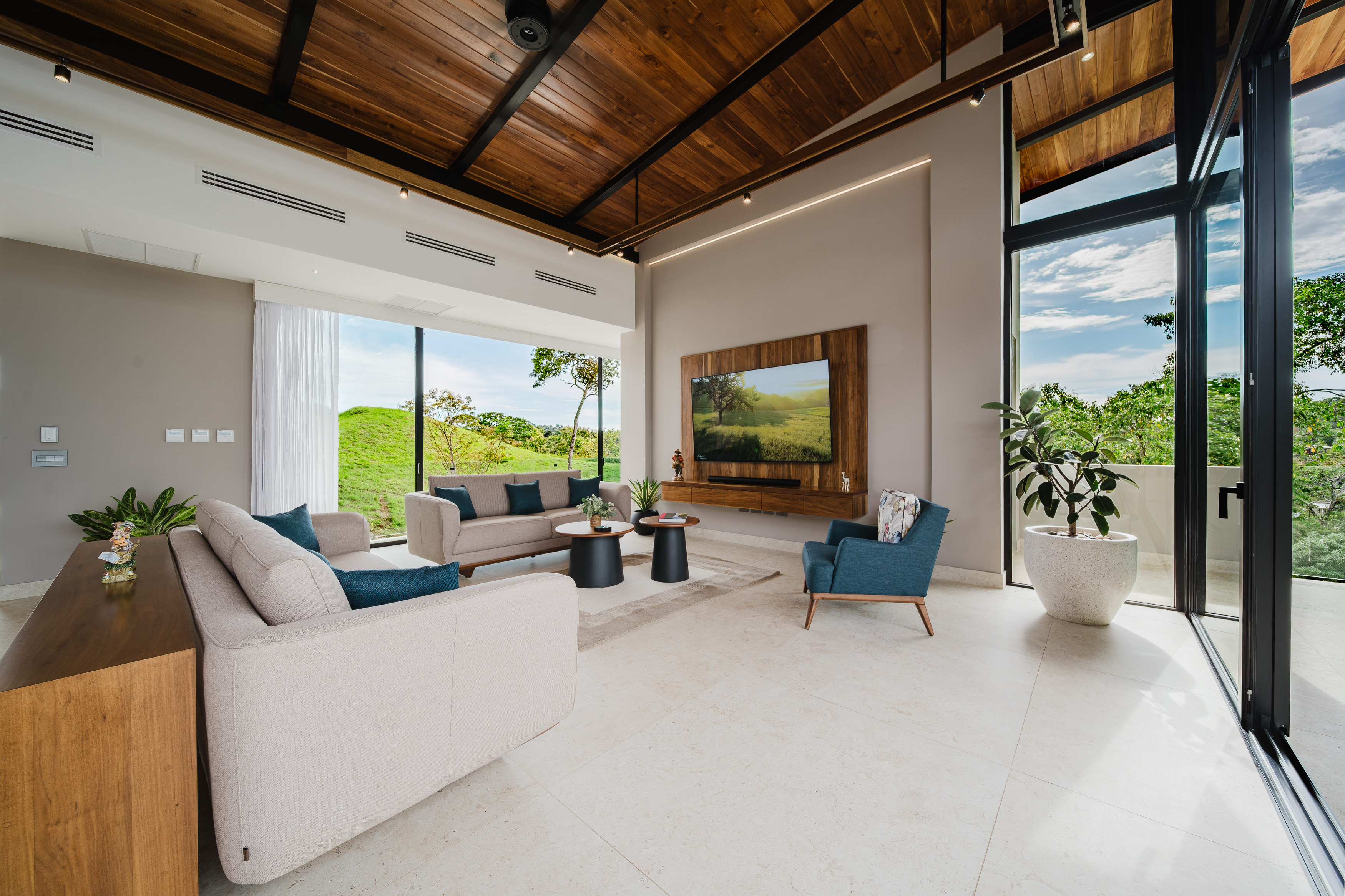 The living room features a high vaulted wooden ceiling, light tiled floors, and expansive glass doors that merge the interior with views of green hills and trees.