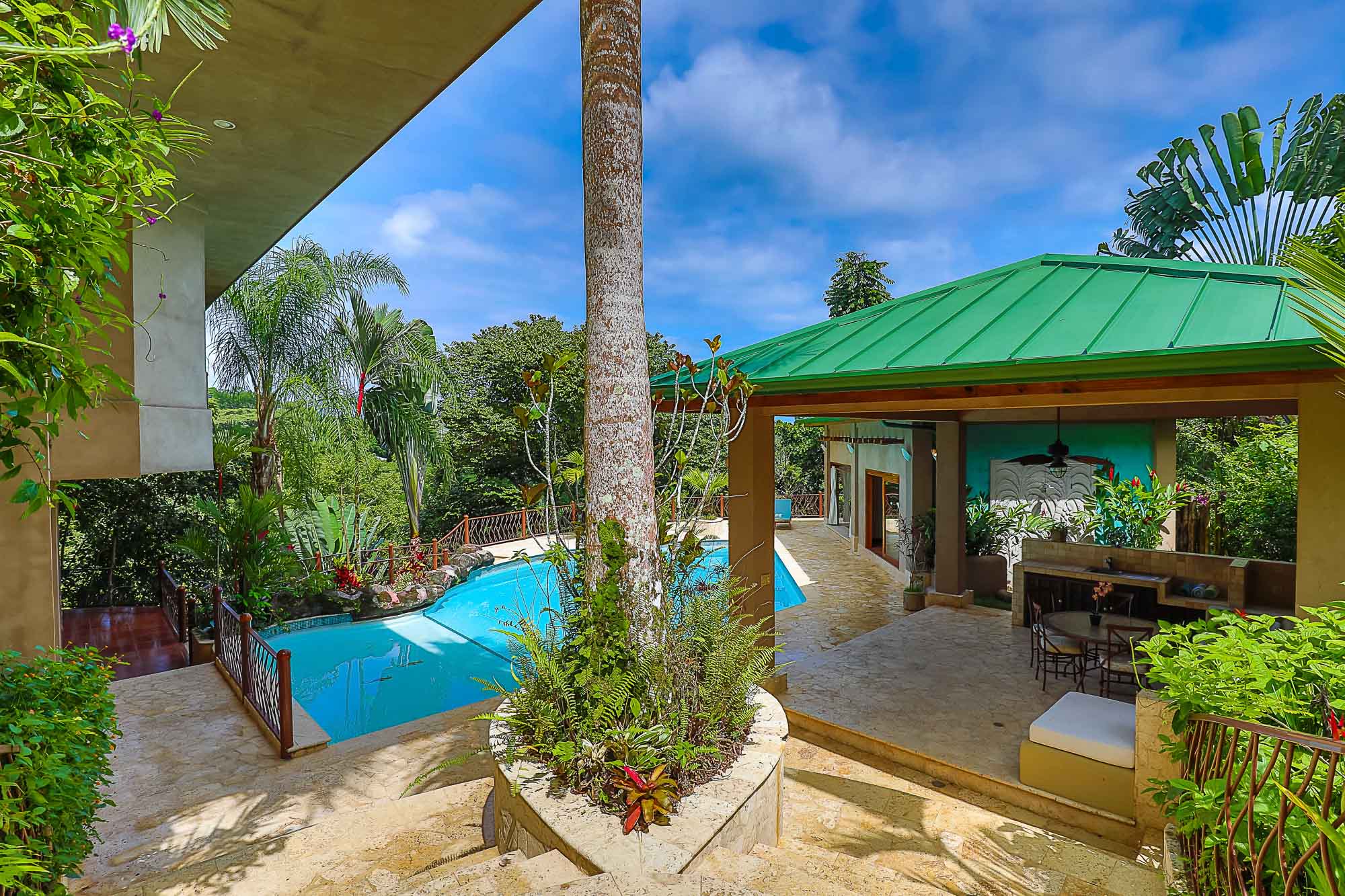 A bright blue pool and a covered outdoor pavilion with an outdoor kitchen and lounge area are nestled within lush tropical foliage under a clear, sunny sky.