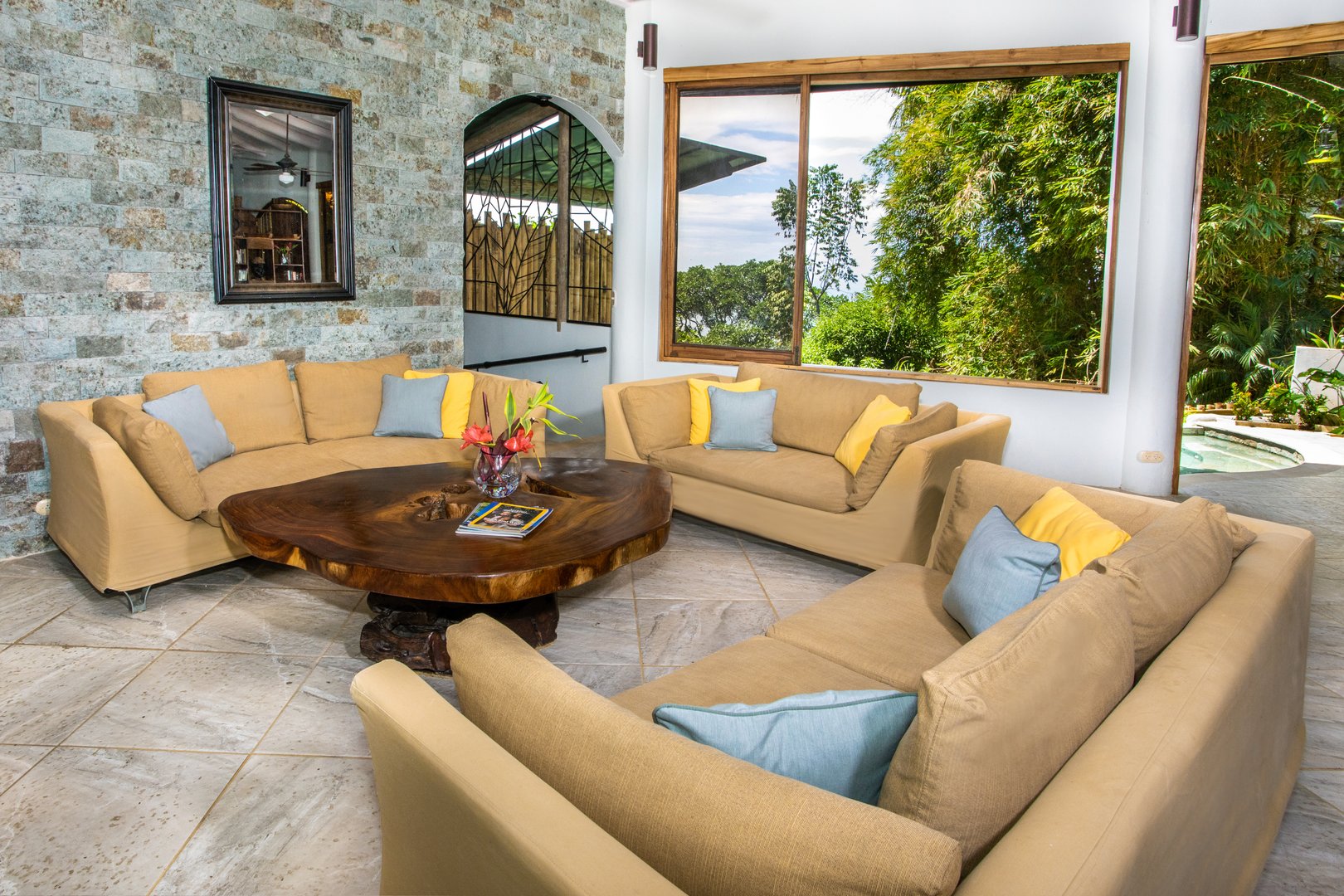 An open living area with a stone feature wall and large windows provides natural light and views of the surrounding jungle, with a pool visible just outside.