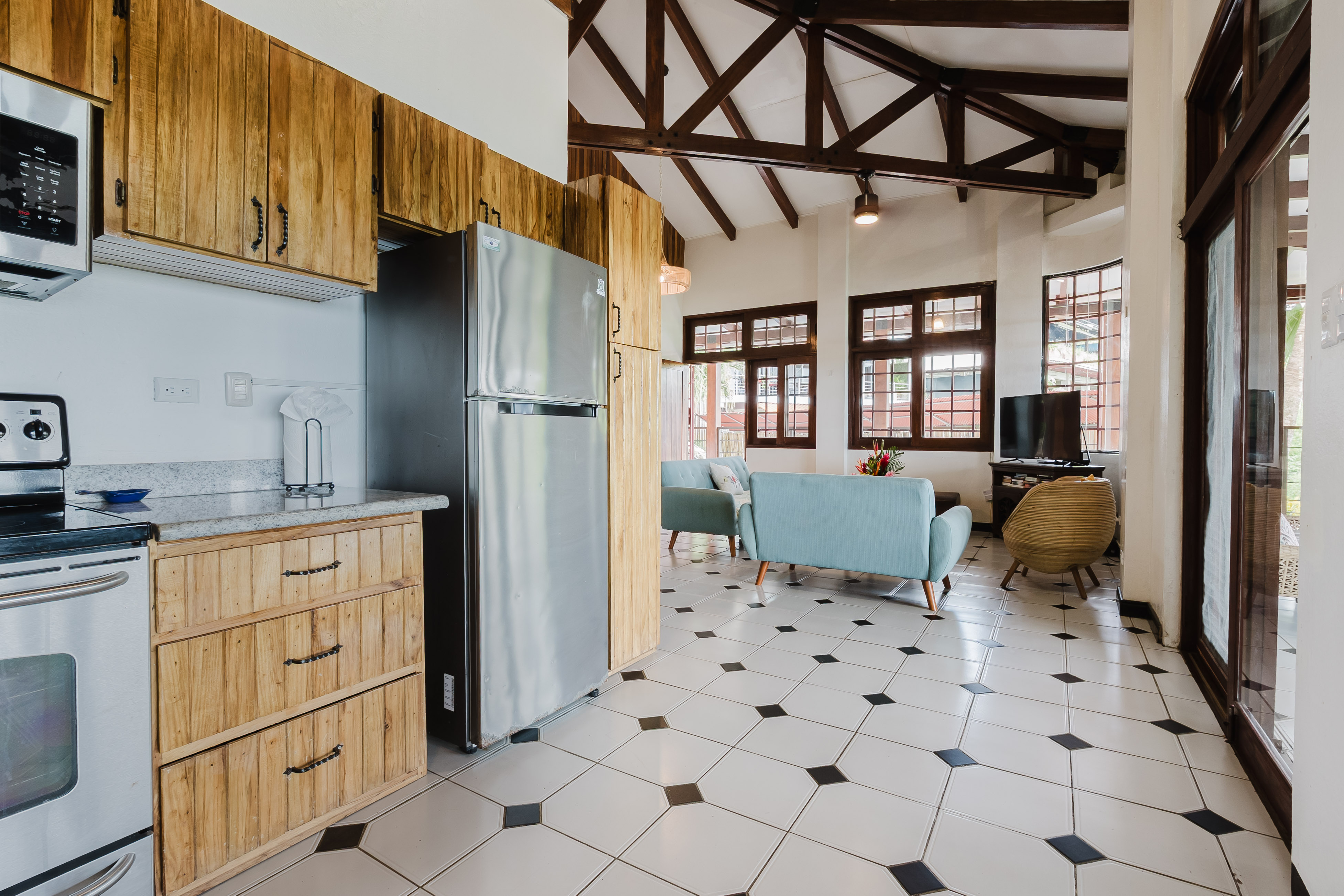 The well-equipped kitchen, featuring natural wood cabinets and stainless steel appliances, opens to a bright living area with exposed beam ceilings, light blue seating, and grid-paned windows providing exterior views.