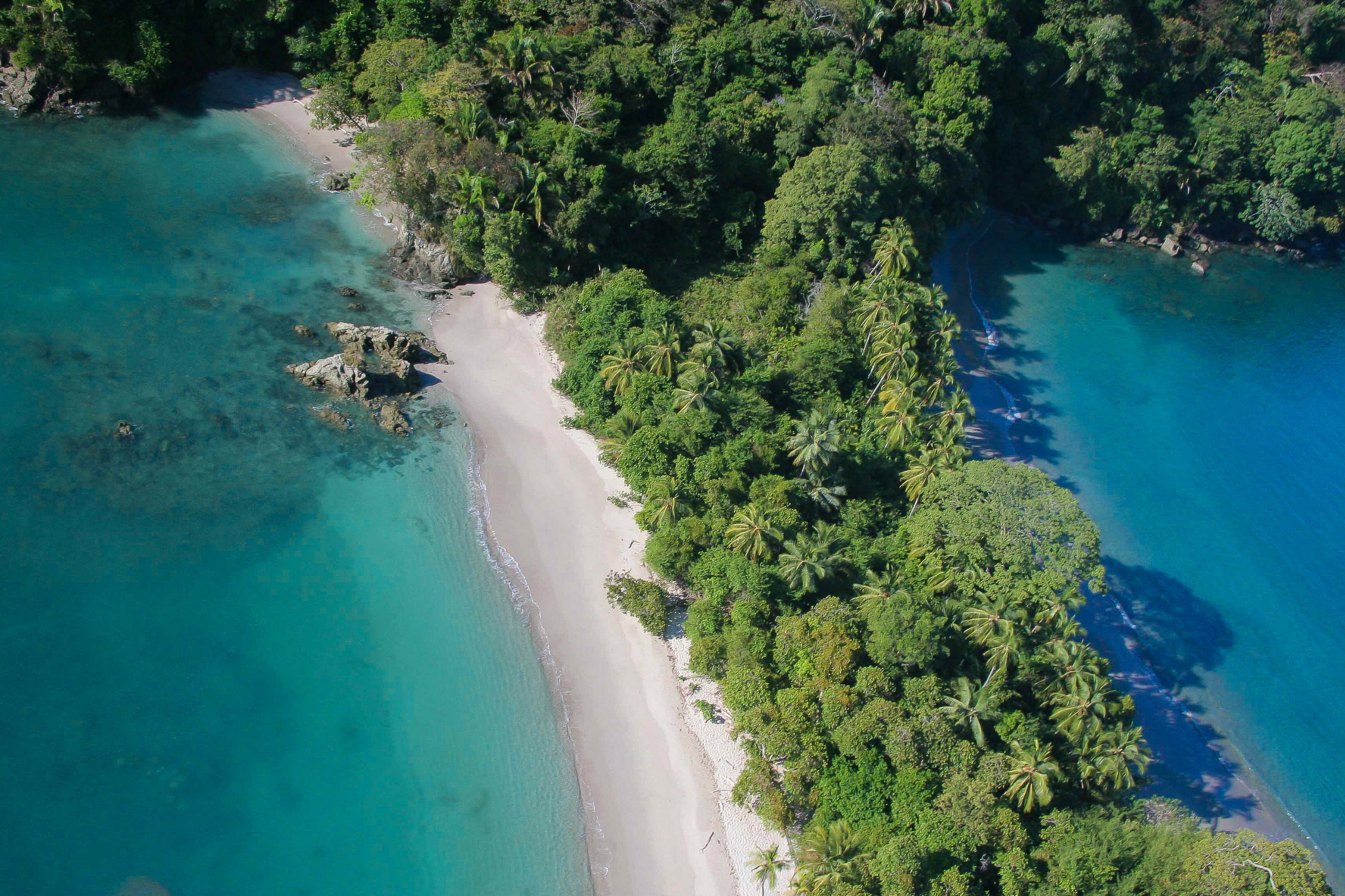 An aerial view reveals a pristine white sand beach curving alongside clear turquoise ocean water, bordered by lush tropical jungle.