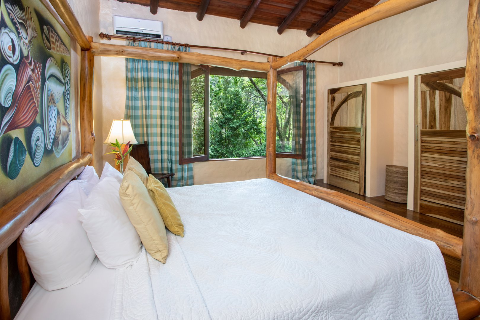 A rustic bedroom features a four-poster wooden bed, exposed ceiling beams, and a large window overlooking dense jungle foliage.