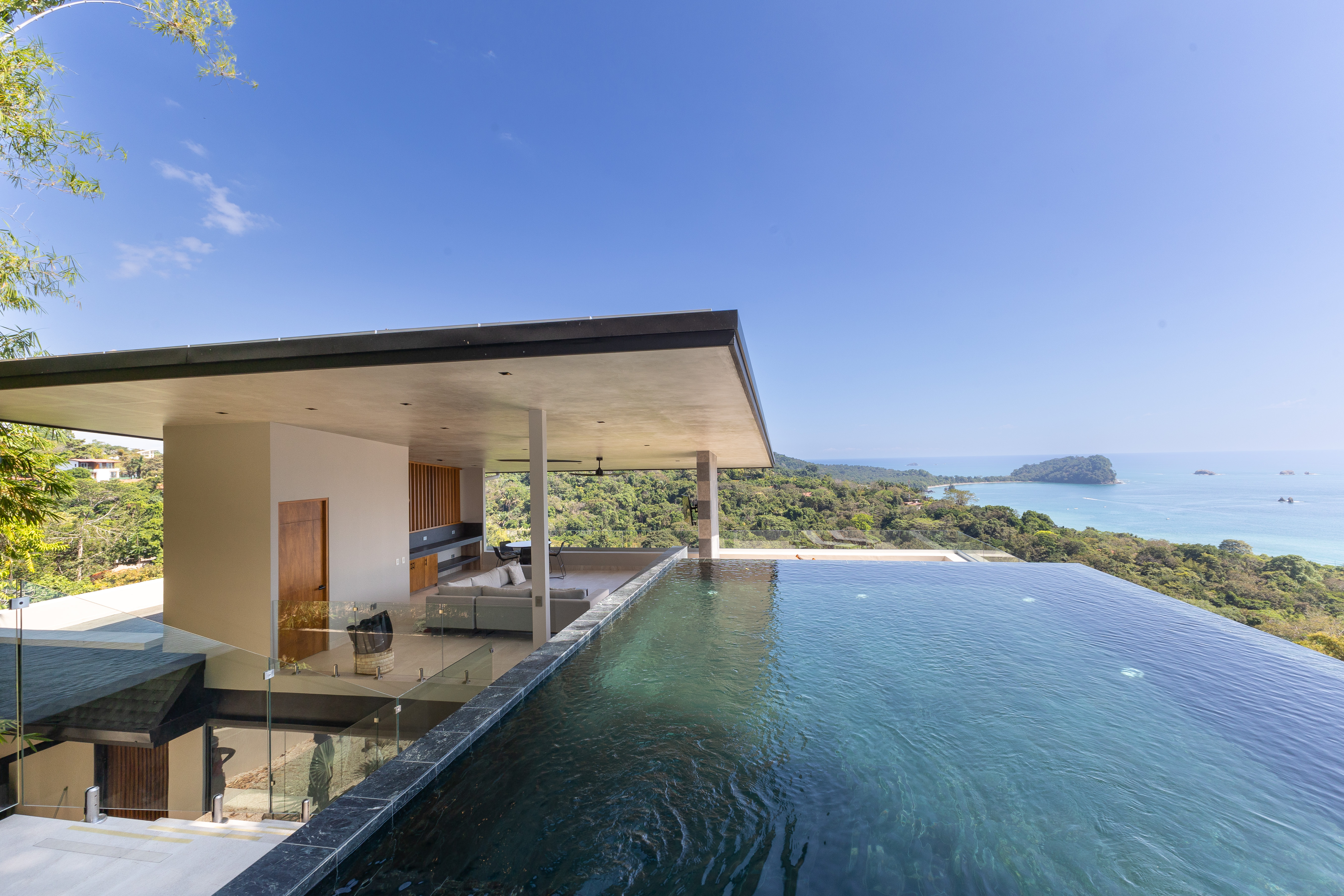 The rooftop infinity pool flows towards expansive ocean and jungle views, complemented by an open-air lounge and outdoor kitchen area under a contemporary shaded structure.