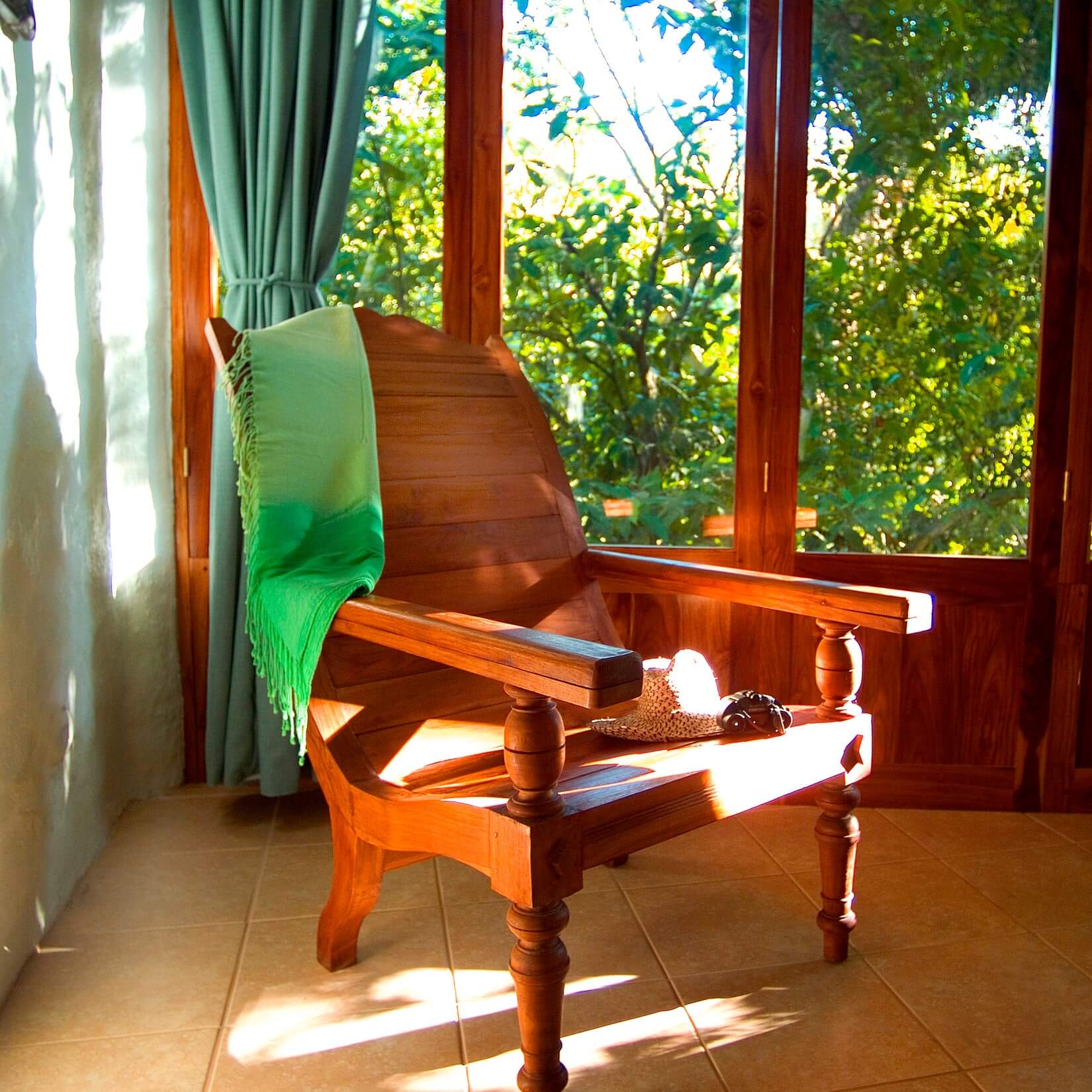 A wooden armchair sits by a large window with dark wood frames, providing abundant natural light and a direct view into the vibrant jungle.