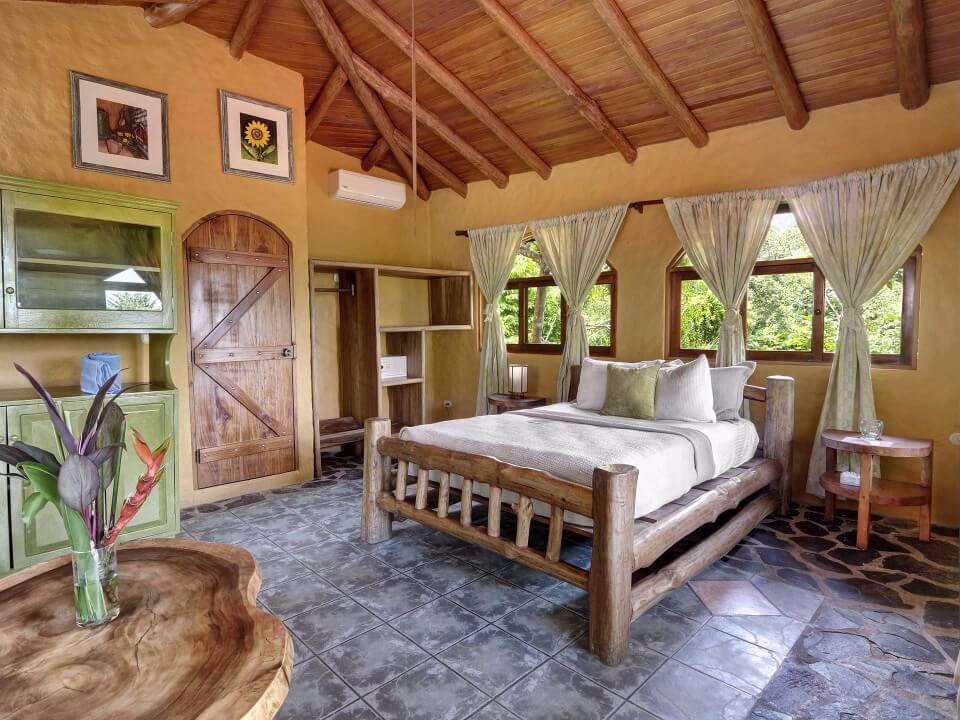 This bright bedroom includes a rustic log bed, a vaulted wooden ceiling with exposed beams, and windows offering abundant natural light and jungle views.