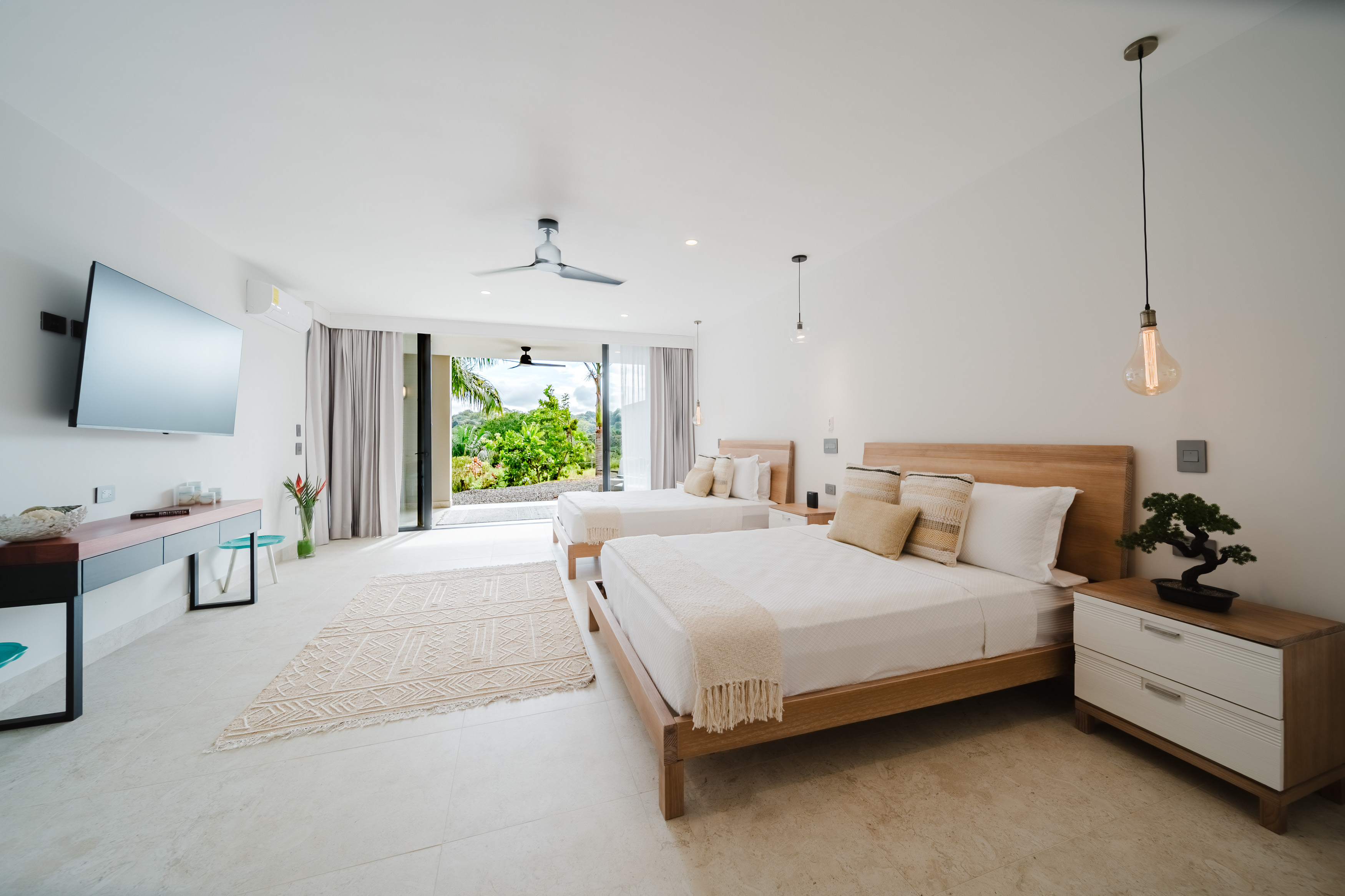 This airy bedroom, with two beds and light-colored floors, opens fully to a vibrant tropical garden through expansive glass doors, inviting the outdoors in.