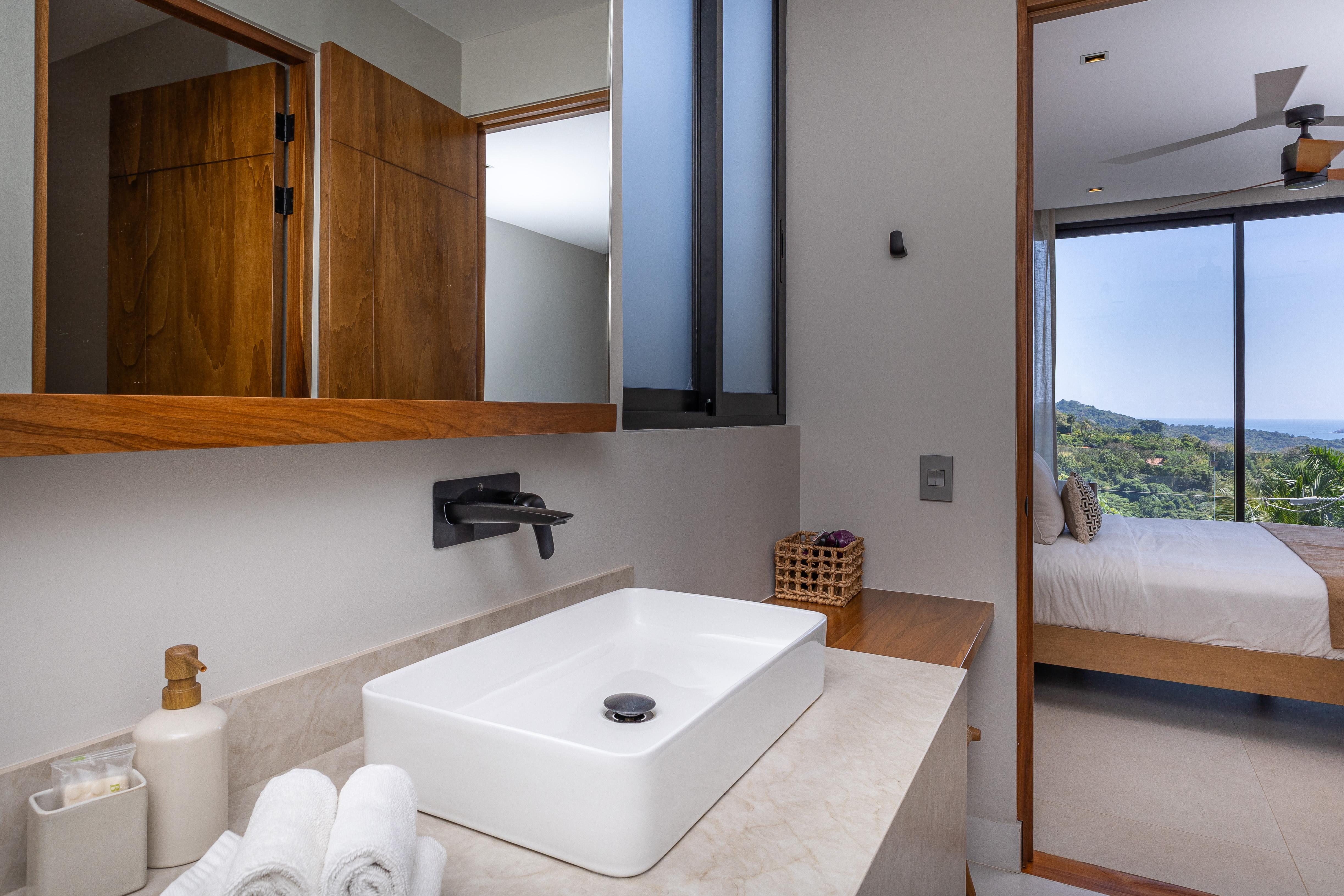 A modern bathroom vanity with a vessel sink and natural wood accents connects to a bright bedroom featuring floor-to-ceiling windows that capture jungle and ocean views.
