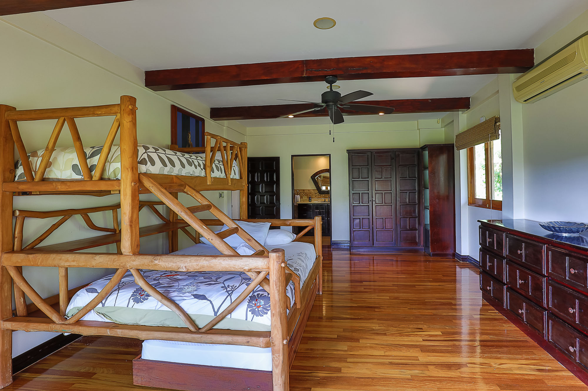 This bedroom offers natural wood bunk beds and a warm wooden floor under exposed ceiling beams, with natural light from a window and an open doorway leading to an en-suite bathroom.