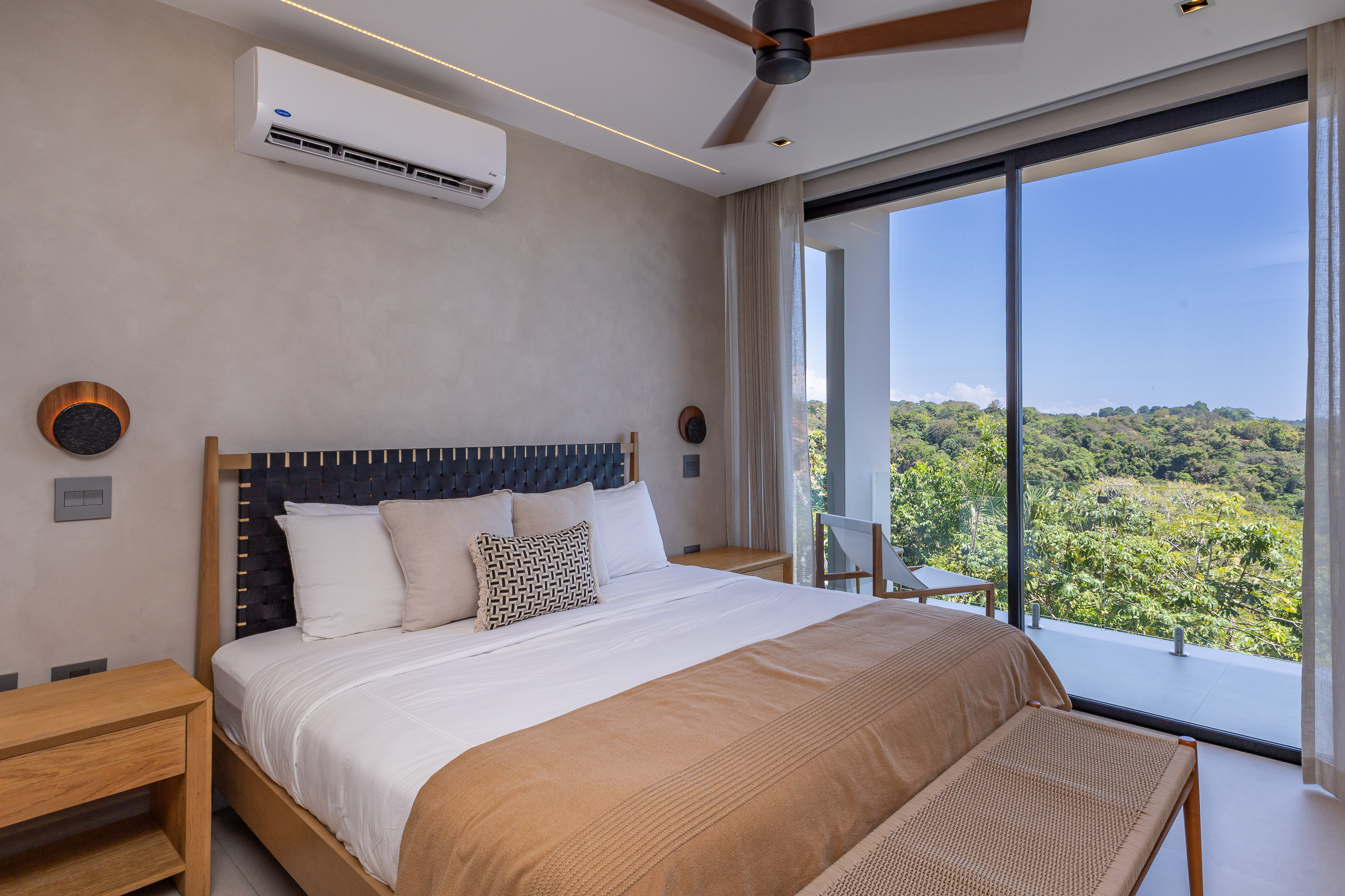 A contemporary bedroom with a king-size bed and modern lighting features floor-to-ceiling glass doors that open to a private terrace and extensive jungle views, inviting natural light into the space.