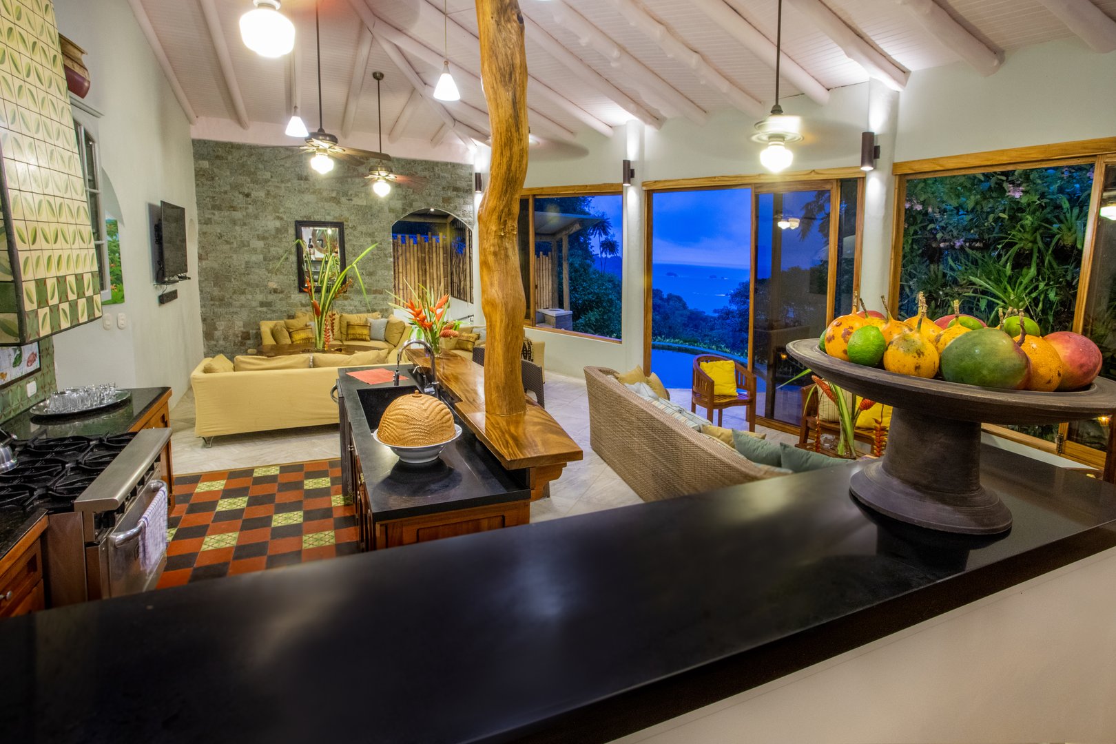 An expansive open-plan kitchen and living area features vaulted ceilings, a stone accent wall, and large windows revealing ocean and jungle views at dusk.