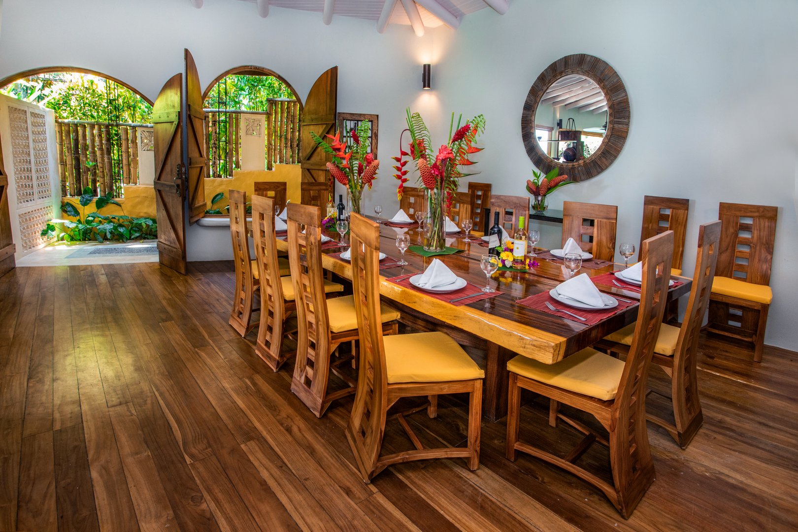 A spacious dining room features a vaulted white ceiling over a dark wood table with seating for ten guests, and arched doorways opening to a tropical garden, inviting natural light.