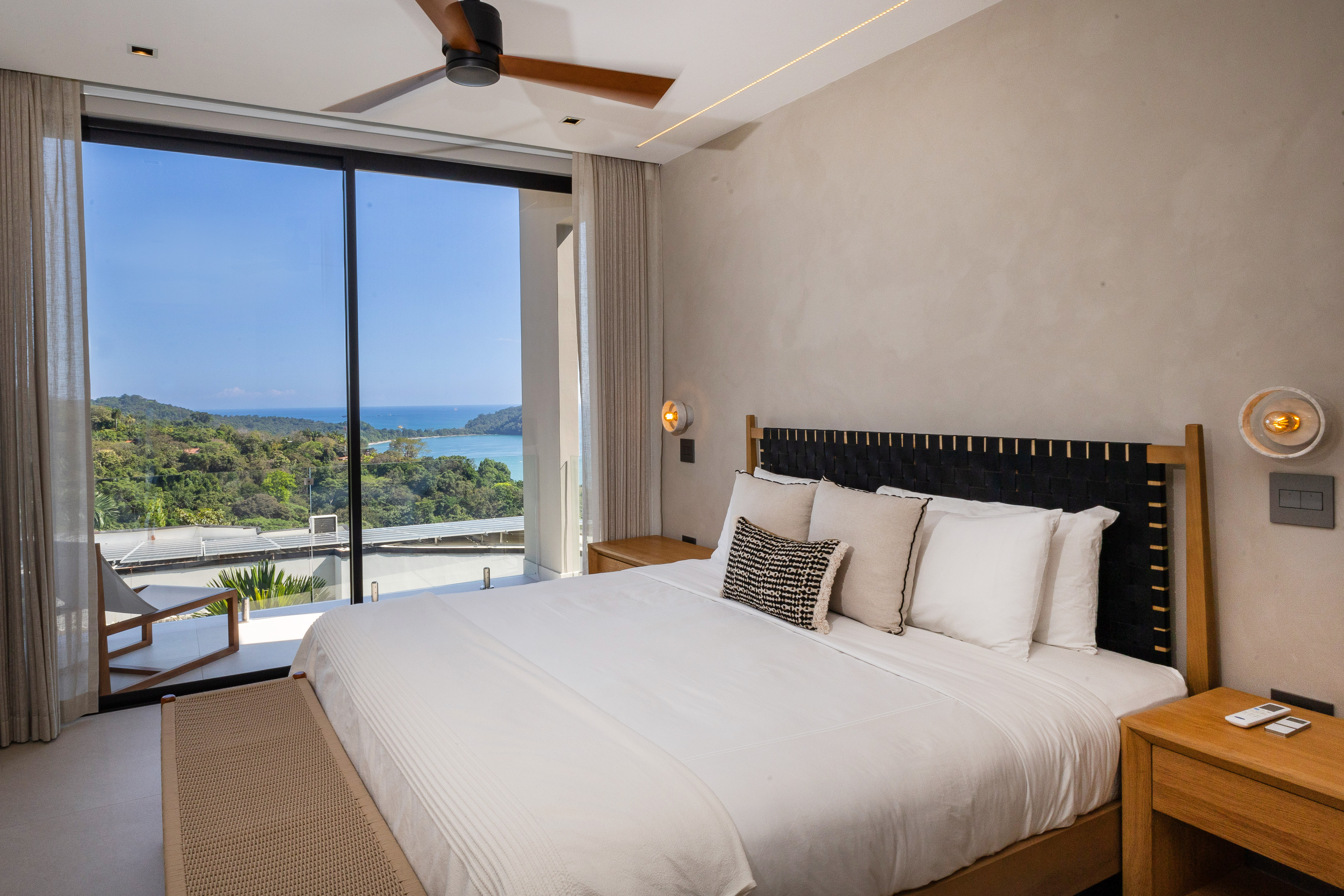 A contemporary bedroom features a large bed, ceiling fan, and floor-to-ceiling glass framing views of the ocean and lush jungle.
