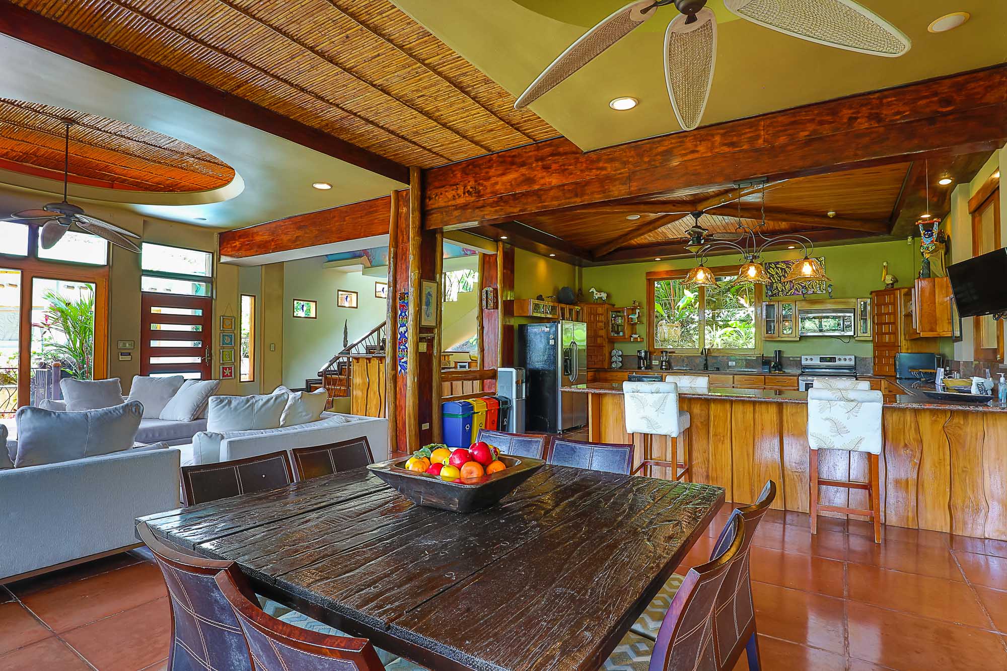 This open-concept main floor combines a comfortable living area with sofas, a large wooden dining table, and a well-appointed kitchen, all featuring natural wood accents, ample light, and views of the lush rainforest.