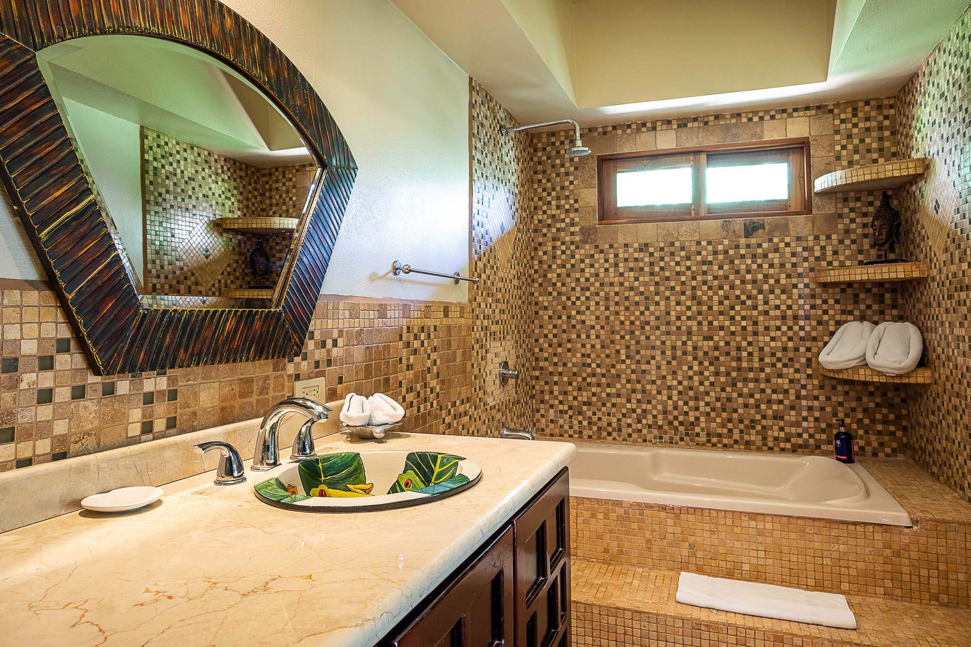A tropical-inspired bathroom features mosaic tile walls, a built-in soaking tub with a rain shower, a marble vanity with a unique leaf-motif sink, and natural light from a horizontal window.